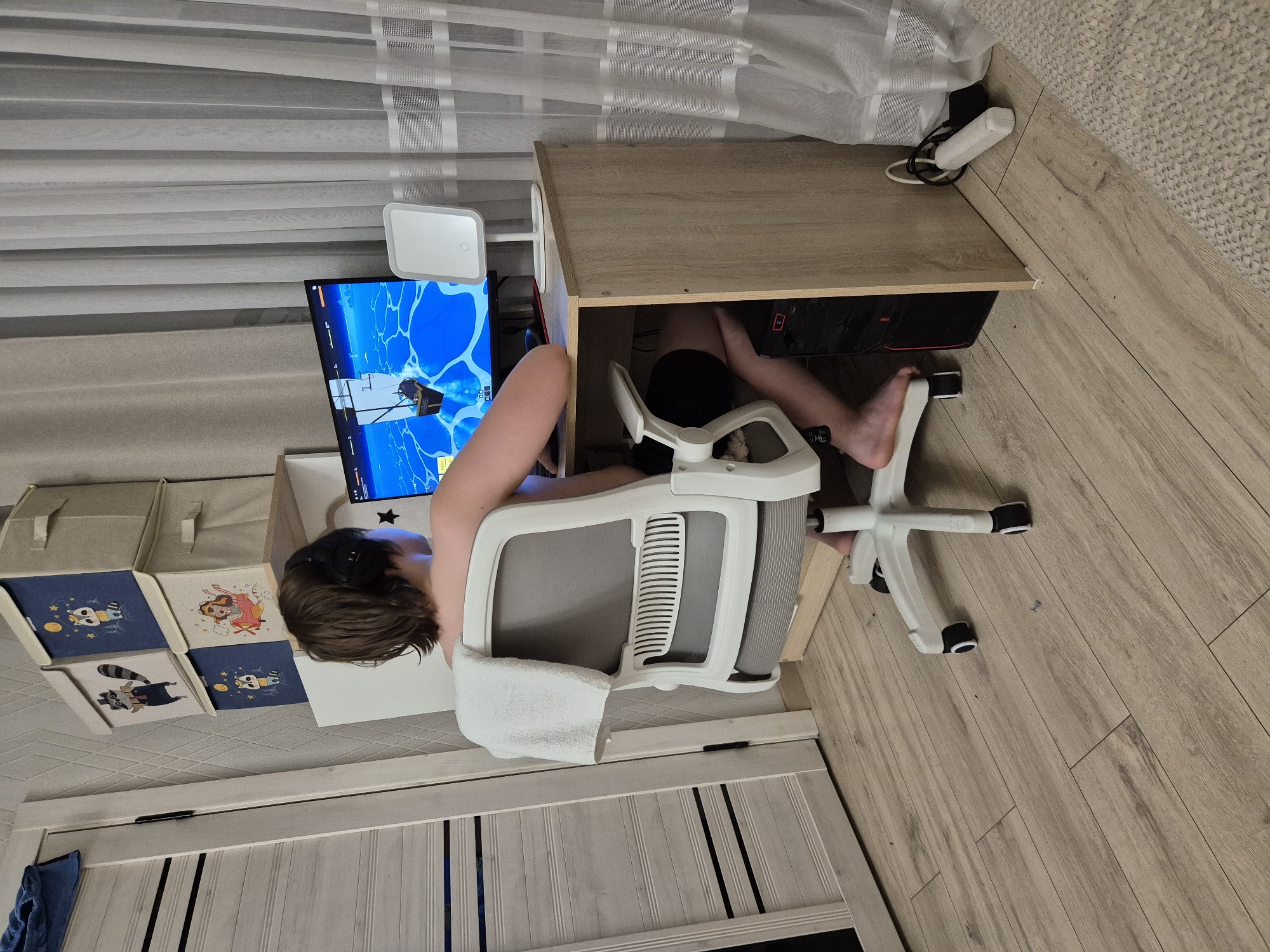 In a room, a young person with headphones is playing a video game on a computer. The person is seated in a white and gray office chair with a towel draped over the back. The computer monitor displays a game with a nautical theme. To the right of the monitor is a small white lamp. A desktop computer tower is visible under the desk. The room has light-colored wooden flooring. In the background, there are three fabric storage boxes with cartoon animal designs stacked on a shelf. A light-colored door is to the left. The scene suggests a typical home environment where someone is engaged in leisure activity. No specific elements in the image provide information about the location being in Astana, Kazakhstan.
