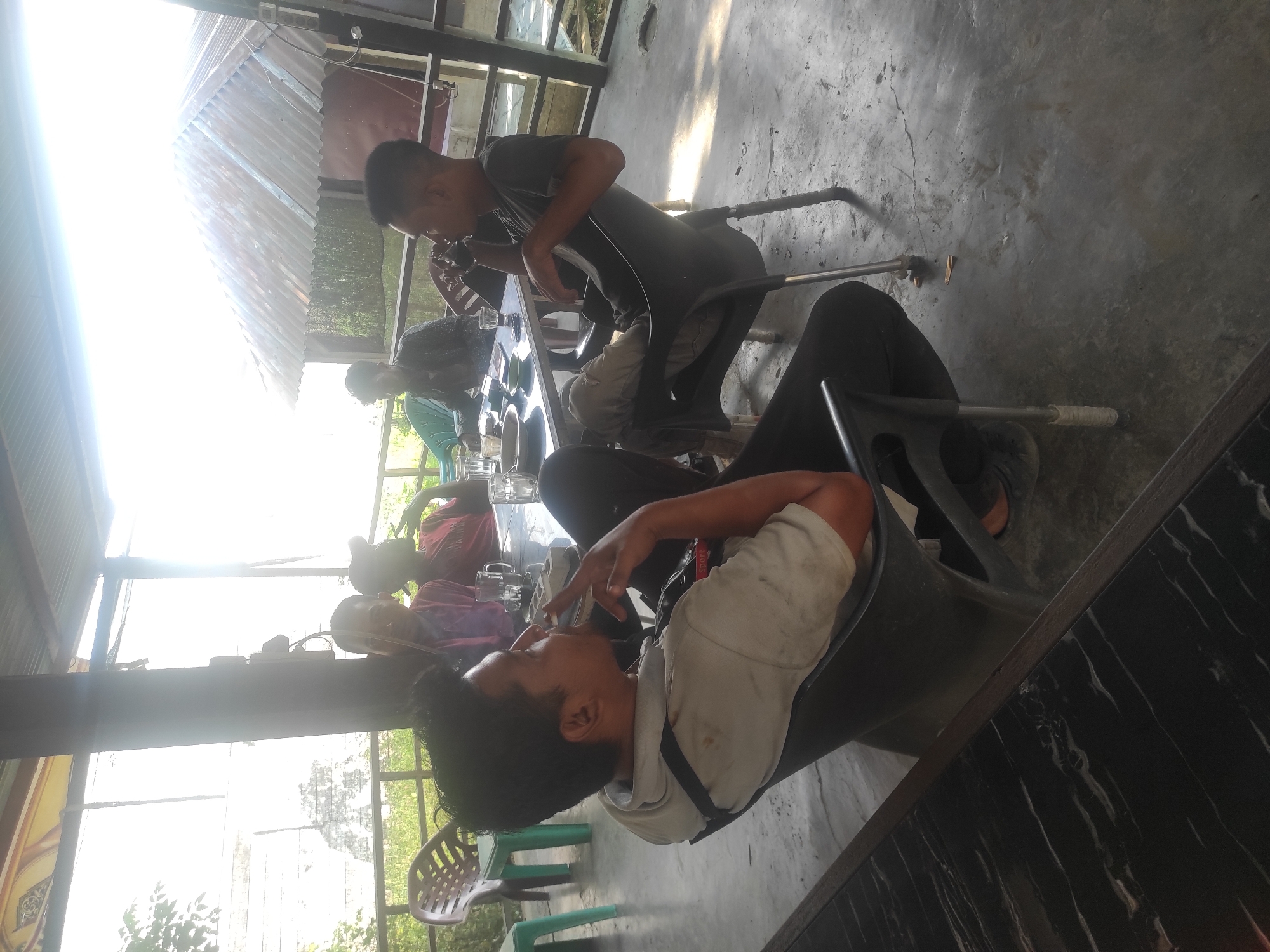 The image shows a group of people at an outdoor cafe in Lhokseumawe, Indonesia. Several individuals are seated around tables. A man in a white shirt is smoking while seated in a black chair. Another man is seated at a table wearing a black shirt. Other people are in the background at other tables. The setting is open-air, with a corrugated metal roof and views of the surrounding vegetation. Glasses and other items are on the tables.
