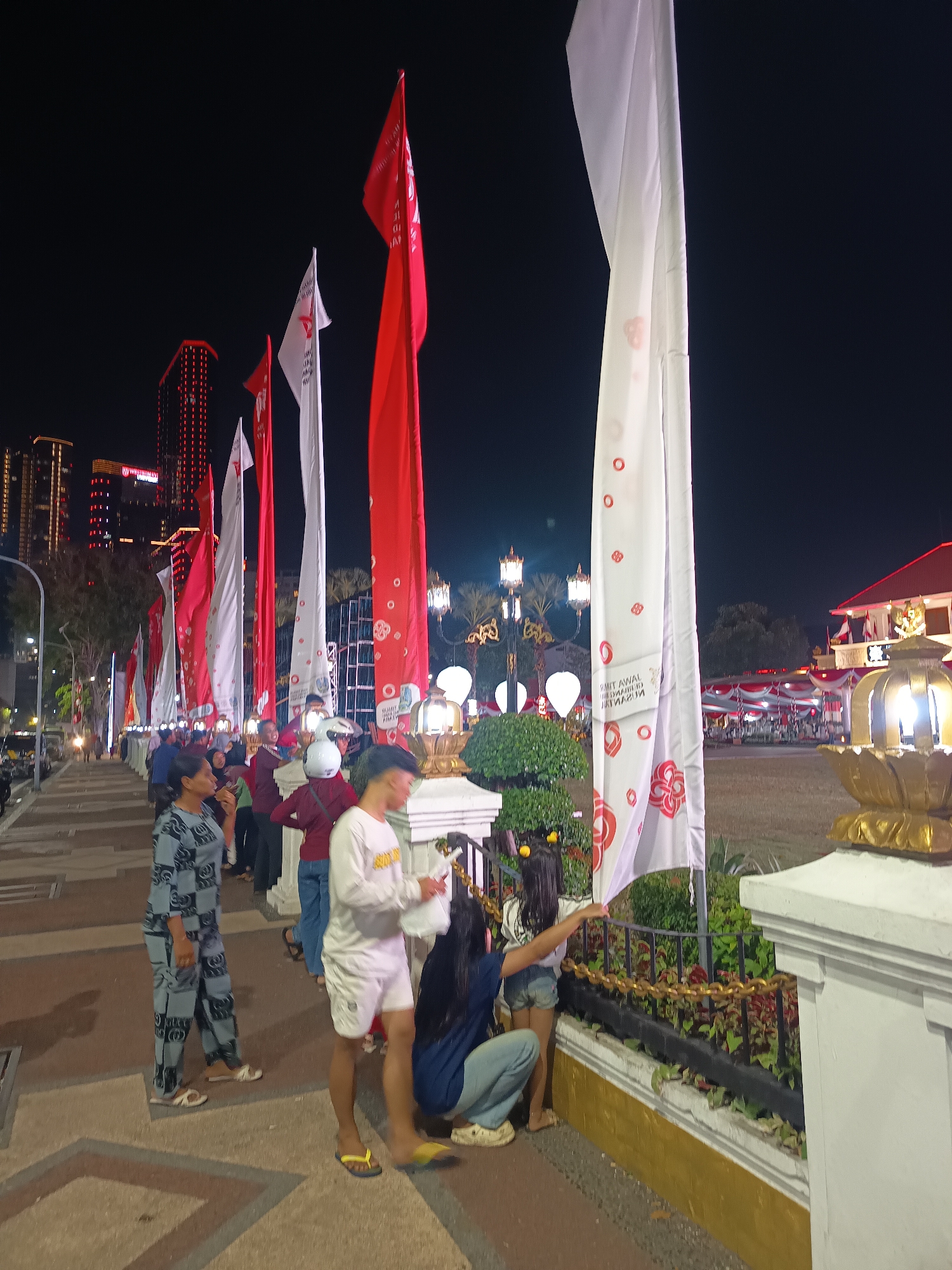 This is a low-angle, nighttime photograph taken in front of the Tugu Pahlawan (Heroes Monument) in Surabaya, Indonesia. The scene depicts a lively outdoor event, likely a celebration or festival, with a crowd of people gathered. Dominating the foreground are several large, vertical flags, predominantly red and white, with some featuring circular patterns and text. In the background, the illuminated Tugu Pahlawan stands tall, its silhouette distinct against the dark sky. The surrounding area is adorned with decorative lights and banners, contributing to the festive atmosphere. People are seen walking along a paved pathway, some interacting with each other, while others are observing the festivities. The image captures a moment of public gathering and celebration in a prominent city landmark.
