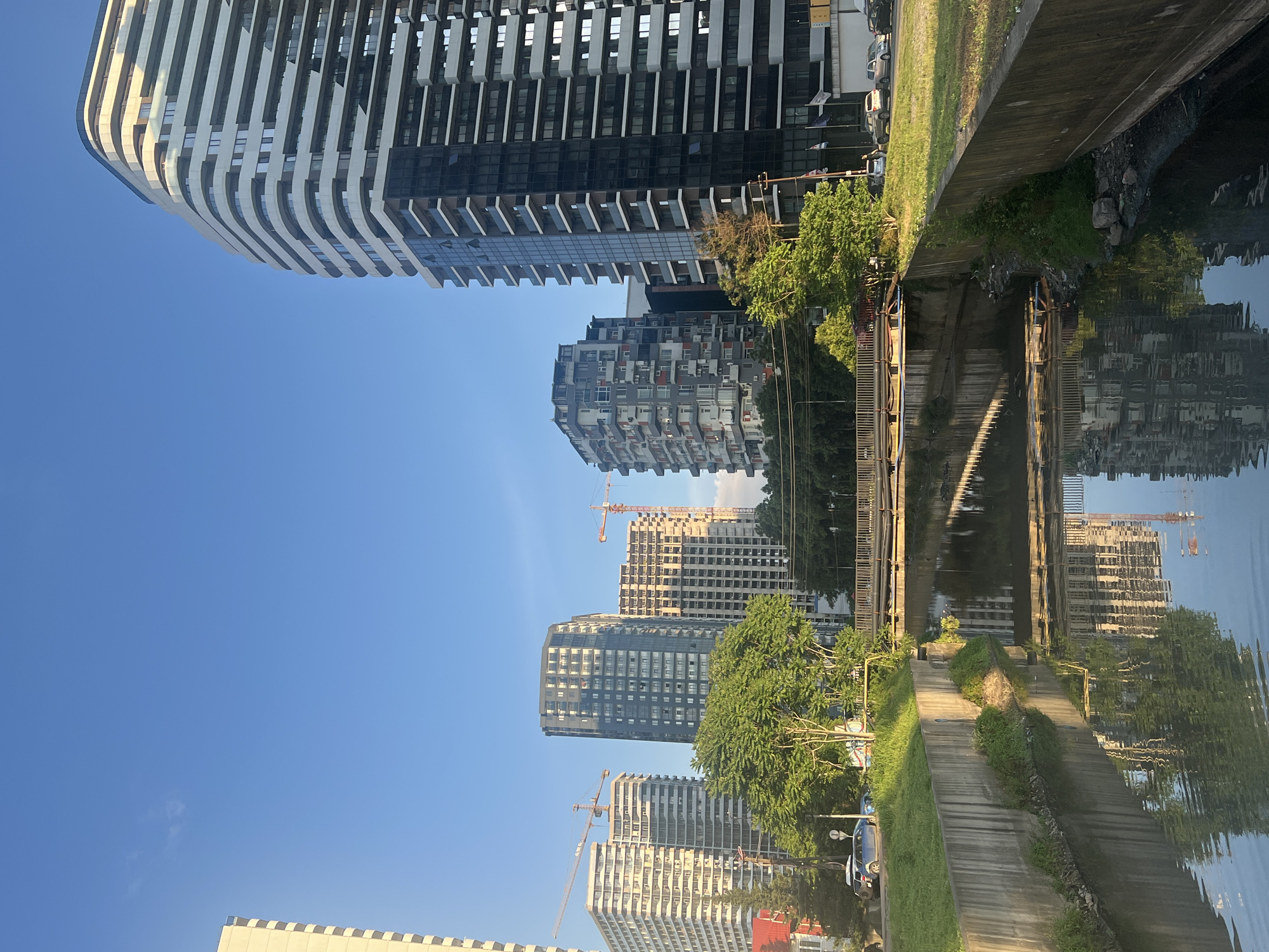 The image depicts an urban canal in Batumi, Georgia, with a focus on modern architecture and a reflection of the cityscape in the water. The canal, bordered by concrete walls, runs horizontally across the lower portion of the frame. A bridge with metal railings spans the canal in the middle ground, supported by pylons. The water's surface is calm, reflecting the surrounding buildings and trees.
On the right side of the canal, a large, contemporary skyscraper dominates the skyline. It features a curved facade with alternating bands of white and glass balconies, giving it a distinctive, layered appearance. To the left of this skyscraper, several other tall buildings rise, some appearing to be under construction, evidenced by cranes visible in the background. These buildings vary in architectural style, with some exhibiting a more angular, block-like design and others featuring numerous balconies and windows.
The left side of the image shows a grassy embankment along the canal, with trees providing a splash of green against the urban backdrop. Cars are parked along the street adjacent to the embankment, suggesting an active city environment. The sky above is clear and blue, with a hint of a wisp of cloud on the far left. The lighting suggests it is either early morning or late afternoon, casting long shadows and highlighting the textures of the buildings.