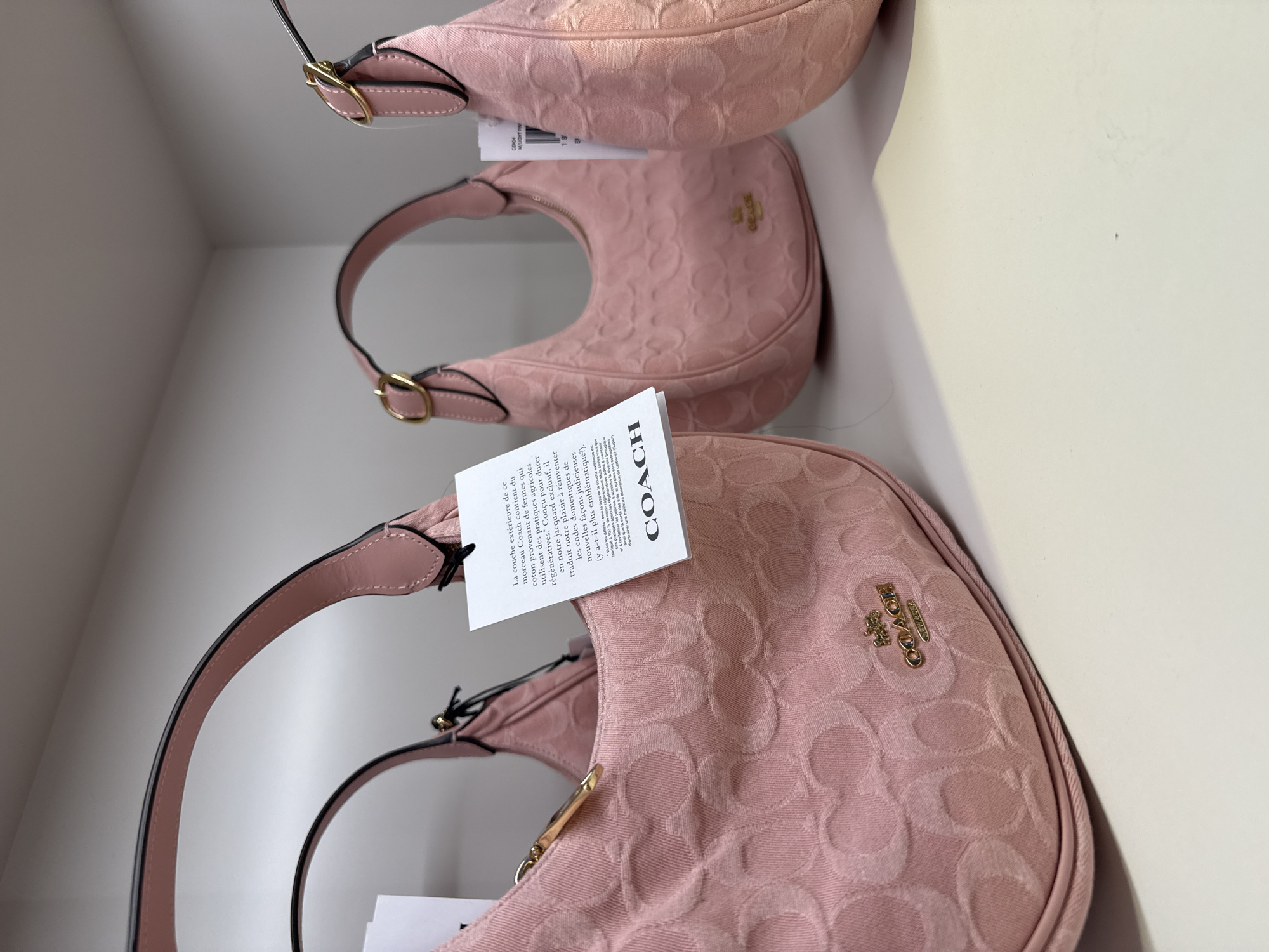 Here is a description of the image:
The image displays three pink Coach handbags, stacked on a shelf. The handbags are the same style, with a curved shape and a short strap. They are made of a fabric with a Coach logo pattern. Each bag has a tag attached. The tags have text in both English and French, including information about the materials used and the brand. The overall setting appears to be a retail environment. The location is in the city of Sunrise, United States of America.