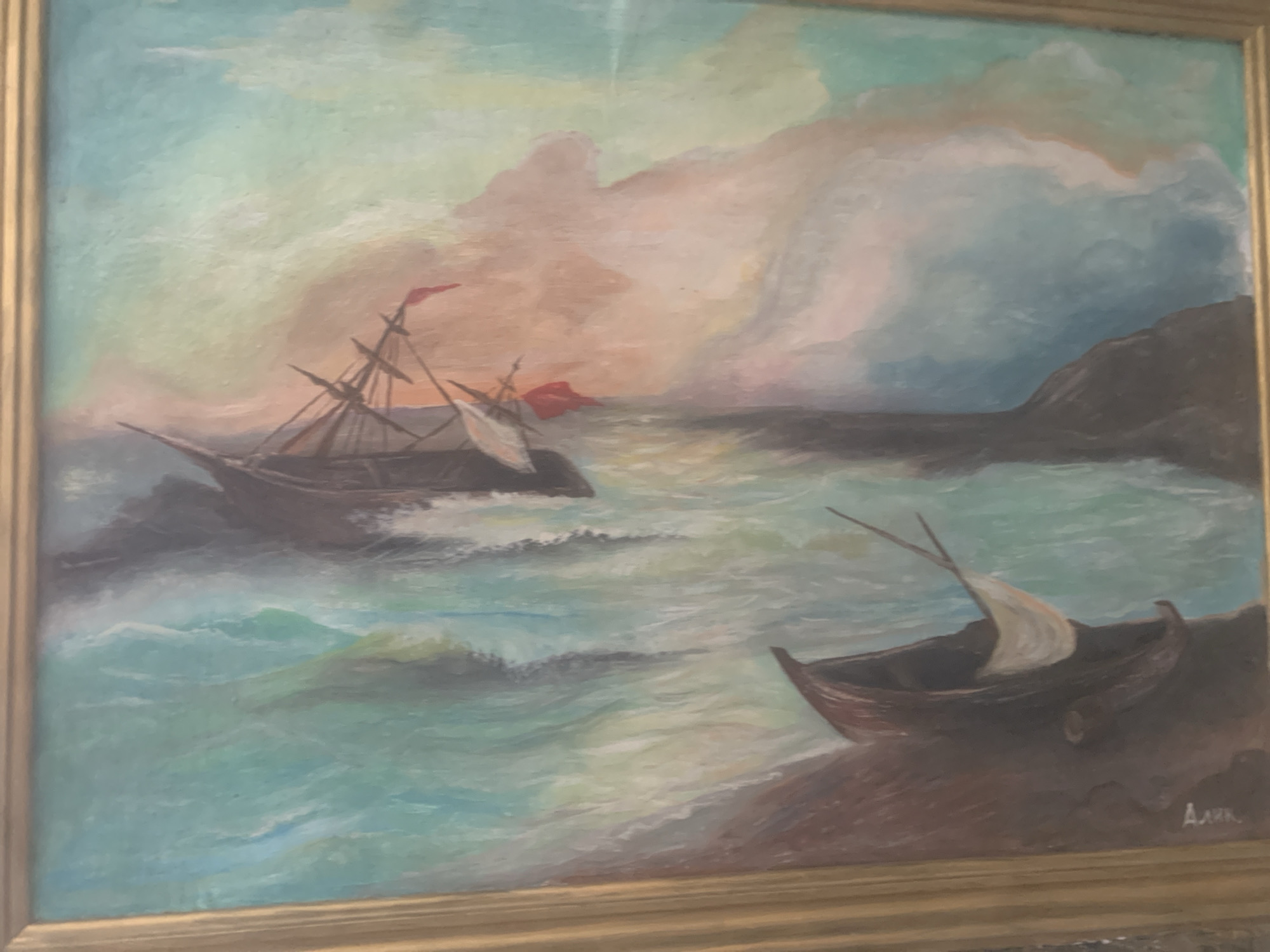 This is a painting depicting a dramatic seascape with two boats. In the foreground, a small, dark boat with a white sail rests on a reddish-brown shore. The water is a turbulent turquoise, with whitecaps and foamy waves. In the background, a larger, dark sailing ship with tattered sails and red flags is partially submerged in the rough sea, listing to one side. The sky is filled with dramatic clouds in shades of orange, pink, and blue, suggesting a stormy sunset or sunrise. The painting is framed by a simple gold frame. There are no people visible in the painting. The signature "Арк" is present in the lower right corner. This painting is not located in the city of Gyumri, Armenia; it is a standalone artwork.