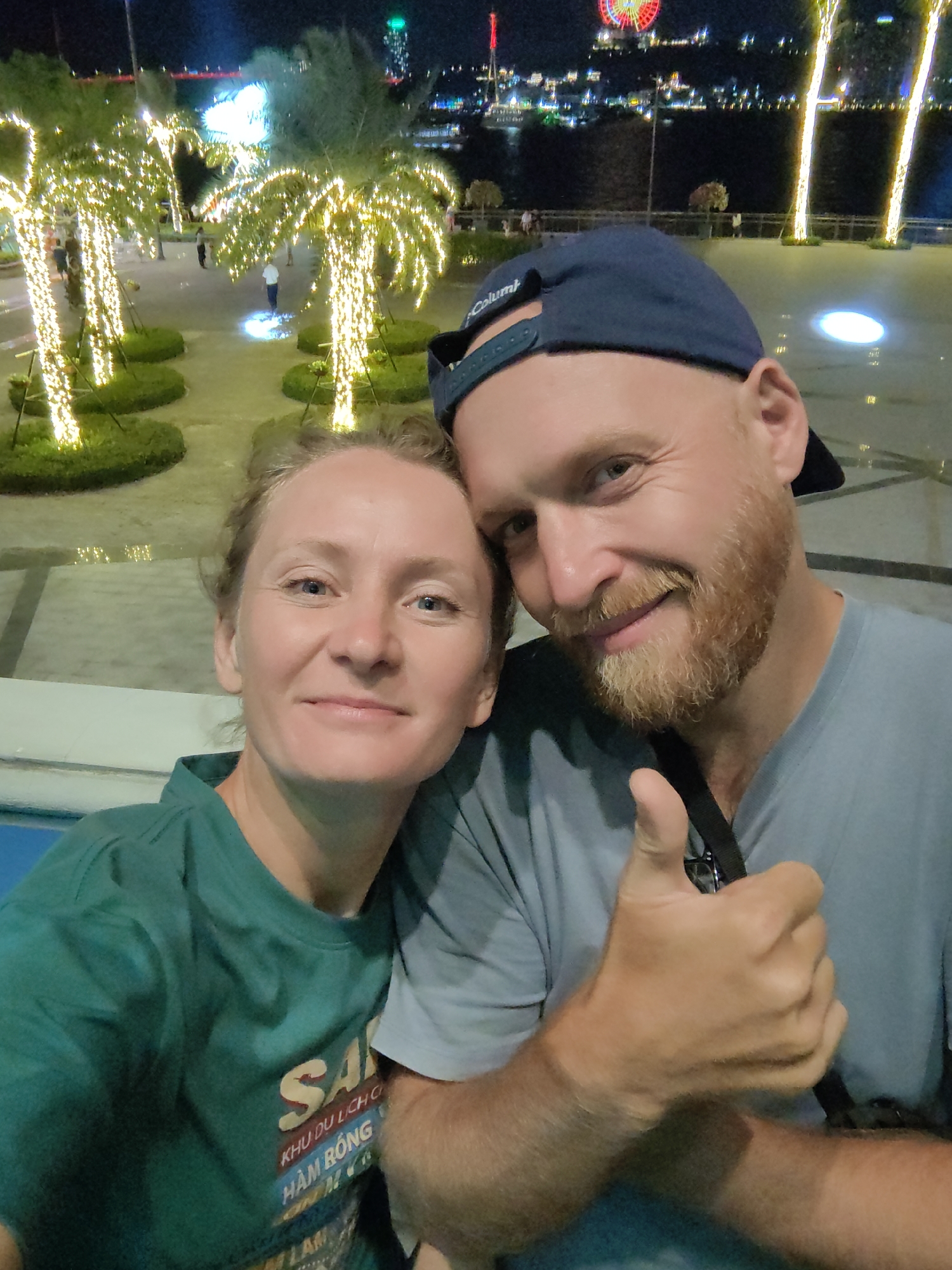 A selfie shows a man and a woman in Bai Chay Ward, Vietnam, at night. The woman, wearing a green t-shirt, stands to the left of the frame. The man, wearing a blue baseball cap and a light blue t-shirt, is on the right and gives a thumbs-up. Behind them, illuminated palm trees line a walkway. In the background, lights mark buildings and a Ferris wheel across a body of water.
