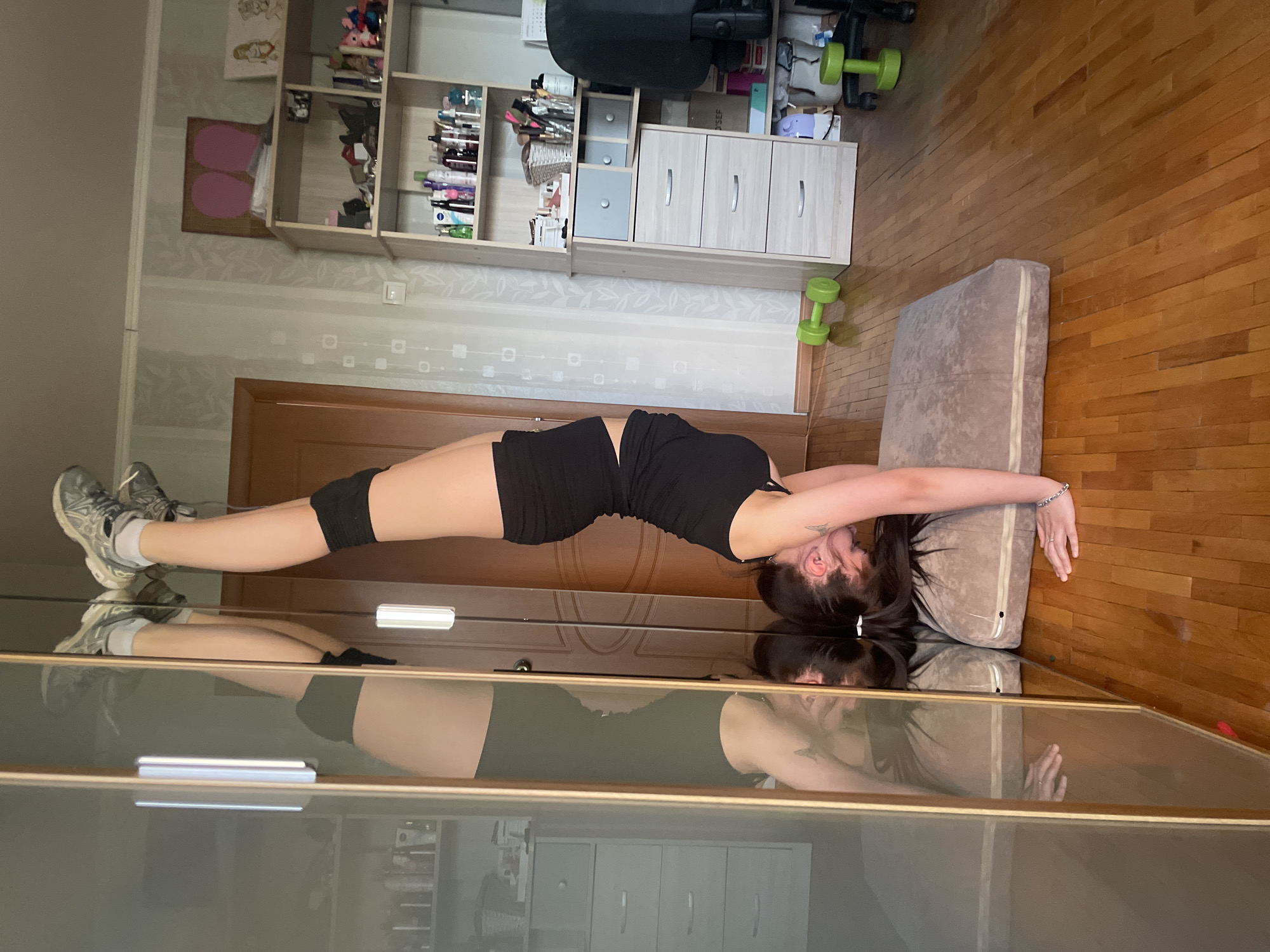 A person in a black tank top and shorts is laying on a tan cushion on a wooden floor. The person's head is on the cushion, and their feet are propped up on a gray shoe cabinet. A reflection of the person and the room can be seen in the cabinet doors. Behind the person, there is a door, shelves filled with various items, and a black office chair. Two green dumbbells are on the floor near the cushion. The location is not identified, and is not necessarily in Saint Petersburg, Russia.