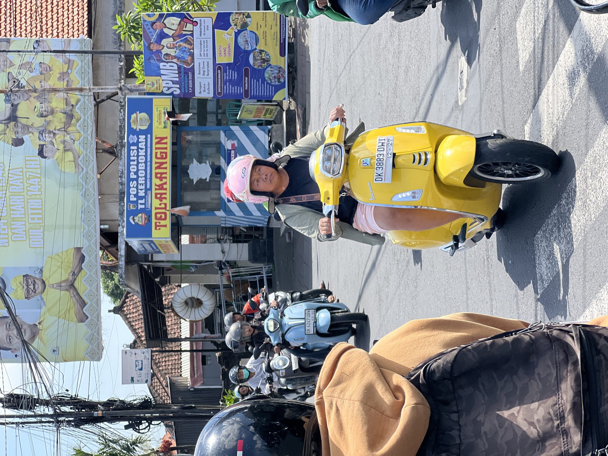 The image depicts a street scene in North Kuta, Indonesia. A person wearing a pink helmet is riding a yellow Vespa scooter. Several other scooters are behind them. Visible signage includes "POS POLISI TL KEROBOKAN" (Police Post TL Kerobokan) and advertisements for "SPMB" (Sistem Penerimaan Murid Baru, or New Student Admission System) for the school year 2026/2027. Power lines crisscross overhead.