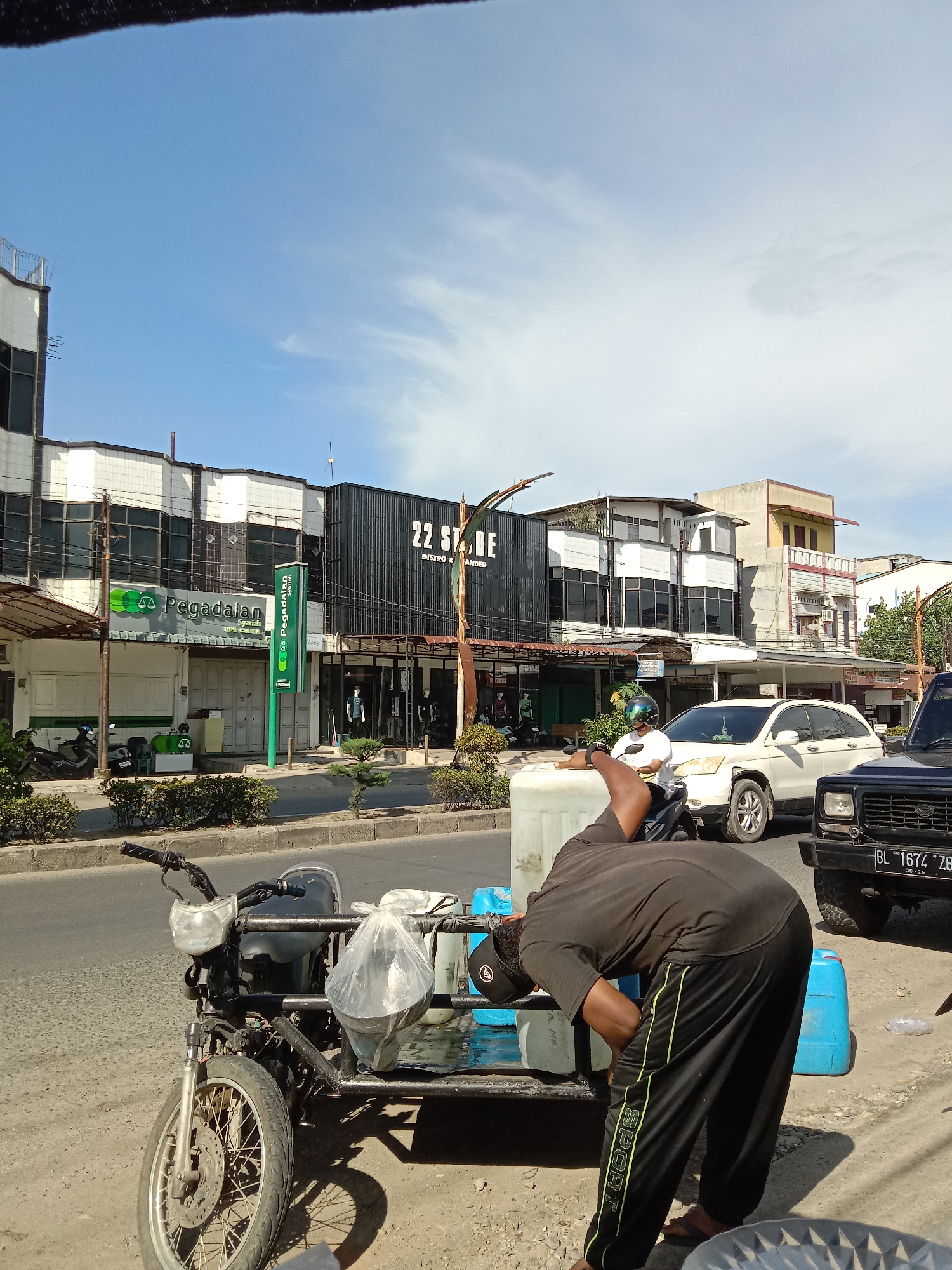 A man is shown working on a motorcycle with a trailer attached, parked on a roadside in an urban area. The motorcycle is carrying several blue containers and a large white container. The man is wearing a black t-shirt and black sweatpants with yellow stripes.
In the background, there is a street with several buildings, including a "22 STRE" store and a "Pegadaian" pawn shop, as well as several cars. The sky is blue with scattered clouds. The license plate "BL 1674 ZB" is visible on a black SUV in the background. The image was taken in Lhokseumawe, Indonesia.