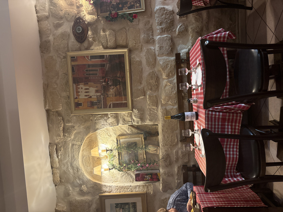 An Italian restaurant's interior features a stone wall with framed artwork and a decorative sconce in a niche. A table, adorned with a red and white checkered tablecloth, is set with glasses and cutlery. A bottle of wine rests on the table. Dark wooden chairs are placed around the table. A partial view of a person sitting at the table is visible.