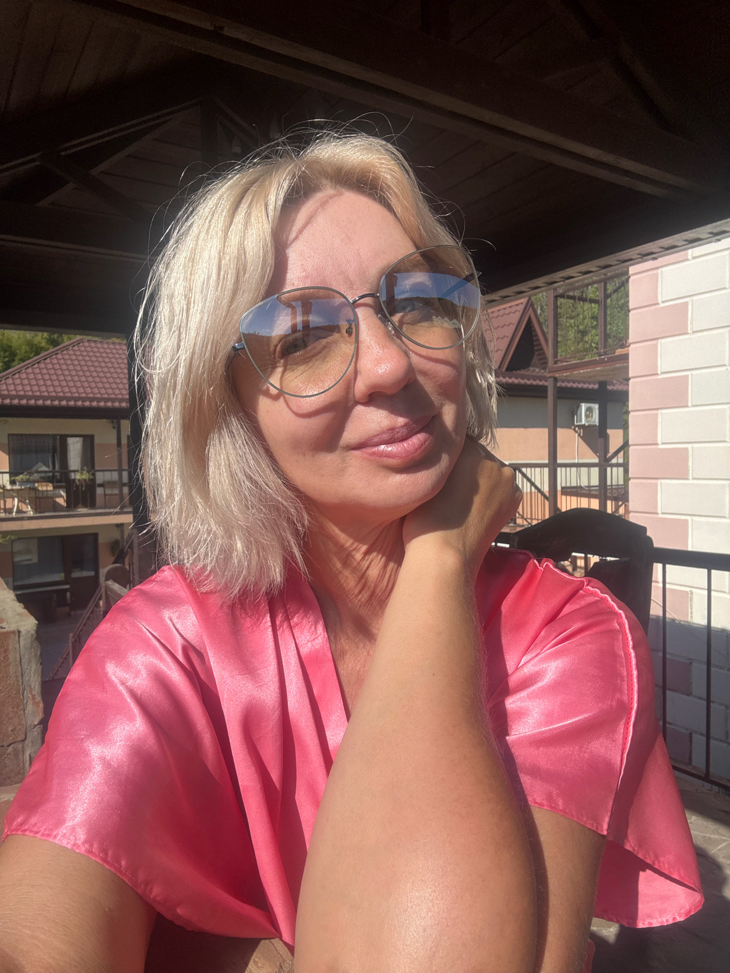 The image is a close-up, selfie-style photograph of a blonde woman wearing large, reflective sunglasses and a shiny pink satin robe. She is outdoors, with her right hand resting on her chin and her left arm visible across the frame. Behind her, there is a building with a red tiled roof and a balcony, along with a light-colored brick structure. The scene appears to be a residential or vacation setting, possibly in a warm climate given the outdoor location and sunlight. The environment suggests a peaceful, relaxed atmosphere. The specific location is a residential area within the resort town of Abrau-Dyurso, Russia, situated on the coast of the Black Sea.