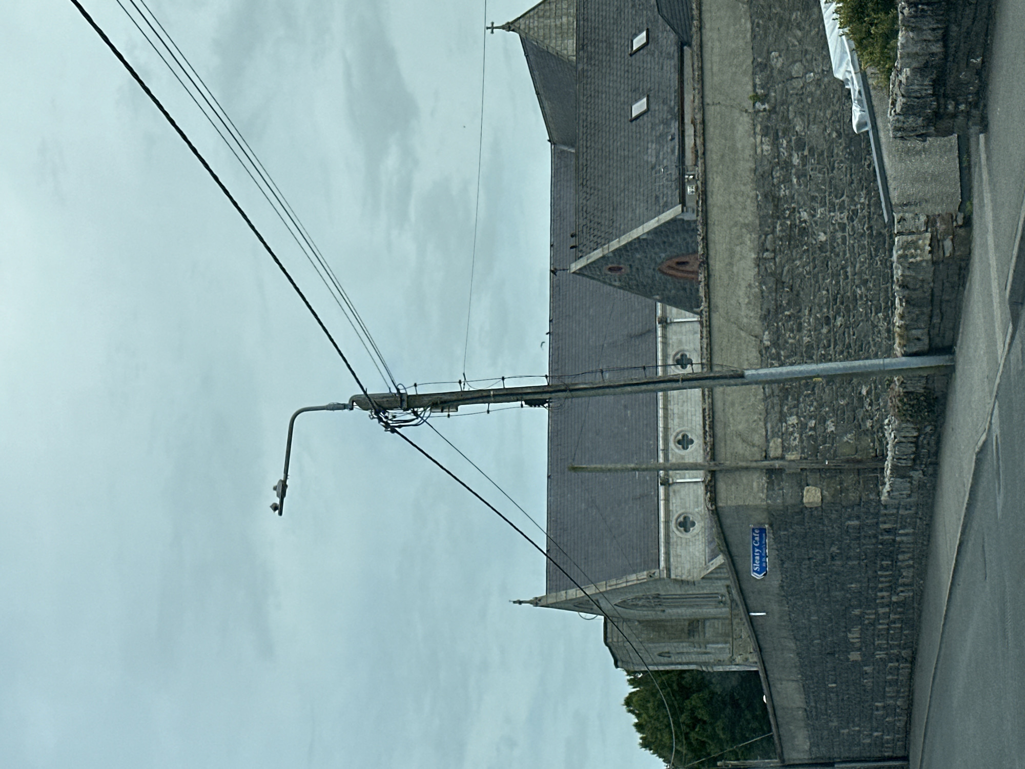 The image captures a cloudy day scene in the city of Carlow, Ireland. Dominating the foreground is a street light with several power lines running across the frame. To the left of the street light, a sign on a stone wall reads "Sleaty Cafe". Behind the wall, the roofline of a church is visible, with a cross atop its spire. The church is constructed of grey stone and features arched windows. Several birds are perched on the roof of the church and on the street light. The overall environment suggests a quiet, possibly overcast, day in a built-up area.