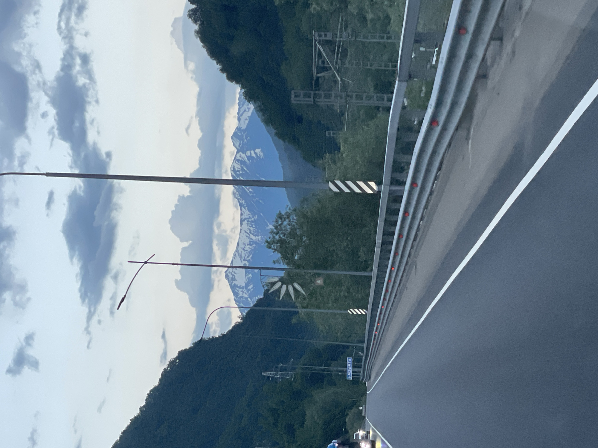 The image shows a road curving through a mountainous landscape, likely at dusk or dawn given the soft lighting and cloudy sky. The road is paved and has a white lane marking. A metal guardrail runs along the side of the road, with red reflectors. On the left side of the road, a steep, tree-covered mountainside rises up. On the right side, there are more trees, overhead power lines, and a distant view of snow-capped mountains. A signpost with blue text is visible on the left side of the road, indicating the location as "р. МЗЫМТА" (River Mzymta). The image appears to be taken from a moving vehicle.