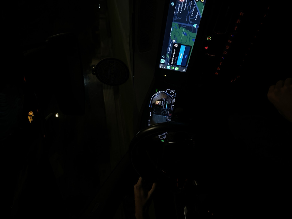 The image depicts the interior of a vehicle at night, viewed from the passenger side, facing the dashboard. A driver's left hand is visible on the steering wheel. A second hand is visible in the lower right corner, likely belonging to a passenger.
The digital instrument cluster behind the steering wheel displays a map segment and "-21.0 C" data. A prominent central infotainment screen shows a navigation application, likely Apple CarPlay, with a map of an urban area. Street names visible on the map include "R. de Mouche", "R. de Mar. Saldanha", and "R. de Pedro de Covilhã". A Portuguese language pop-up on the navigation screen asks, "Conduzir até casa?" (Drive home?), offering "SIM" (Yes) and "NÃO" (No) options, with a route estimated at "7 min - 1.6 km." Below the infotainment screen, vehicle controls with red and orange indicators are partially visible. A circular, dark air freshener or ornament, possibly with "SCX" text, hangs from the rearview mirror area. An illuminated "PASSENGER AIR BAG OFF" indicator is visible on the upper dashboard. The external environment is dark, with minimal ambient light suggesting night conditions.
Based on the visible street names on the navigation display ("R. de Mouche", "R. de Mar. Saldanha"), the vehicle is located in the city of Porto, Portugal, specifically within the civil parish of Aldoar, Foz do Douro e Nevogilde.