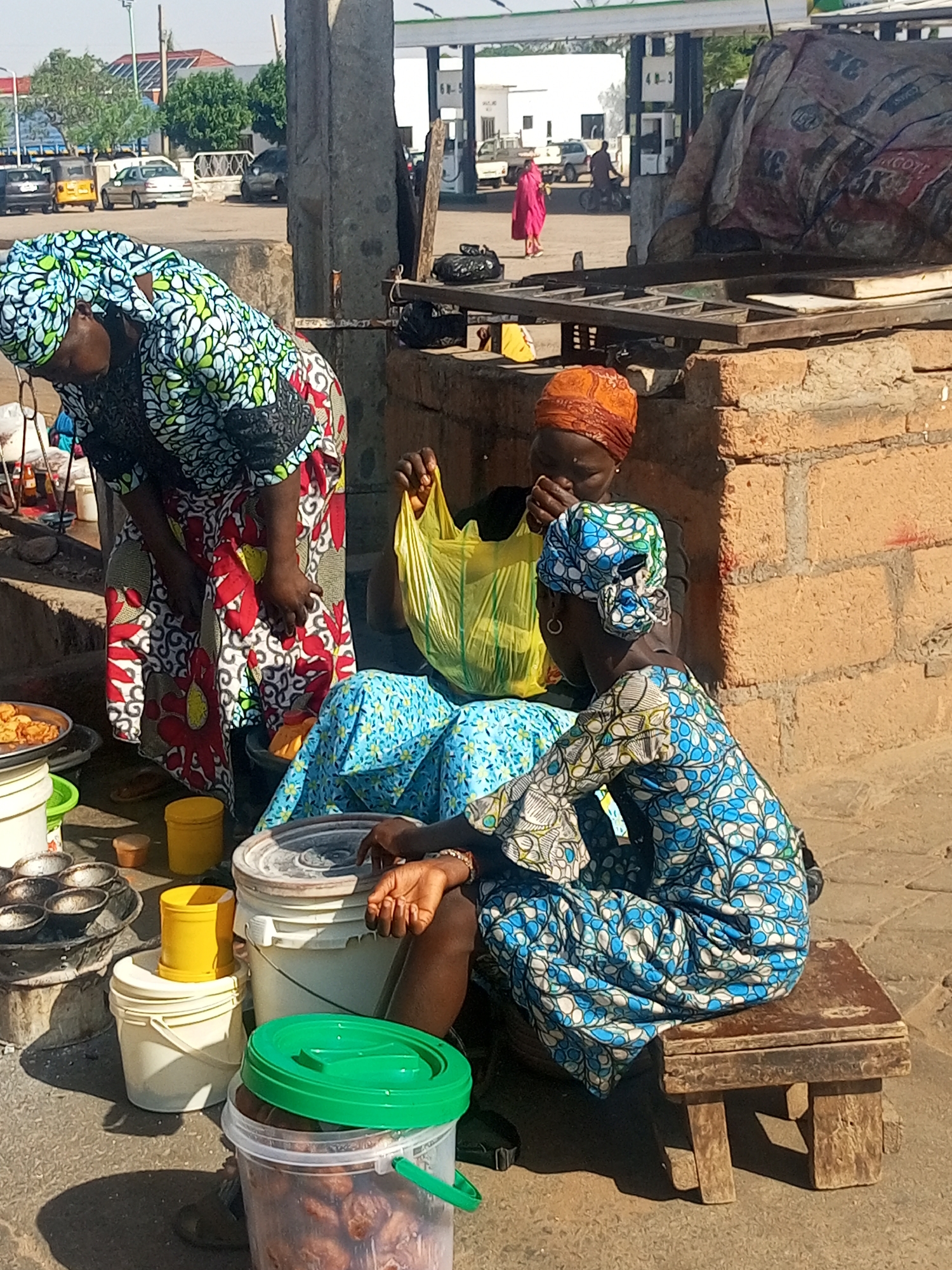 The image depicts three women at what appears to be a vibrant roadside market or food stall in Tempure, Nigeria, under bright daylight.
On the left, a woman in a colorful headwrap and dress, patterned with green, white, blue, and red motifs, is bending over. She seems to be attending to various containers and metal pans, possibly used for preparing or serving food, arranged on the paved ground.
In the center, another woman is seated, wearing an orange headwrap and a dark top. She holds a bright yellow plastic bag, looking down into it as if bagging items or inspecting contents. To her right, a third woman sits on a small, rustic wooden stool. She is dressed in a blue and white patterned headwrap and matching attire, and her hands rest near a large white bucket. She appears to be observing the activity around her.
The foreground is filled with assorted items including multiple plastic buckets of various colors (white, yellow, clear with a green lid containing round, dark food items) and small metal molds, likely for cooking. Short shadows suggest it is midday or early afternoon.
Behind the women, a low brick structure provides a backdrop. Further in the background, a busy street scene unfolds with cars, a person in a bright pink outfit walking, and the canopy and pumps of a gas station. Visible text on the gas pumps includes "6N-5" and "4B-3." A large bundle of sacks, marked with "3X," rests on a metal frame behind the brick structure, possibly containing supplies for the stall. The scene conveys a sense of daily commerce and community activity.