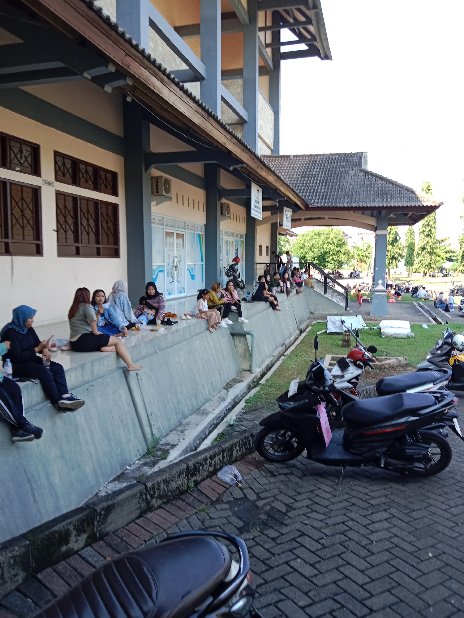 The image depicts an outdoor scene in the city of Kendal, Indonesia, likely in a public area such as a government building or waiting area, possibly the Kendal Regent's Office based on the architectural style and the presence of people waiting. In the foreground, several parked motorcycles are visible on a brick-paved ground. To the left, a group of women are seated on a raised concrete ledge. Some are looking at their phones, while others are conversing. Beyond them, the facade of a building with windows and air conditioning units is visible. Further in the background, more people are scattered around, some sitting on the ground and others standing. The environment suggests a warm, sunny day.
