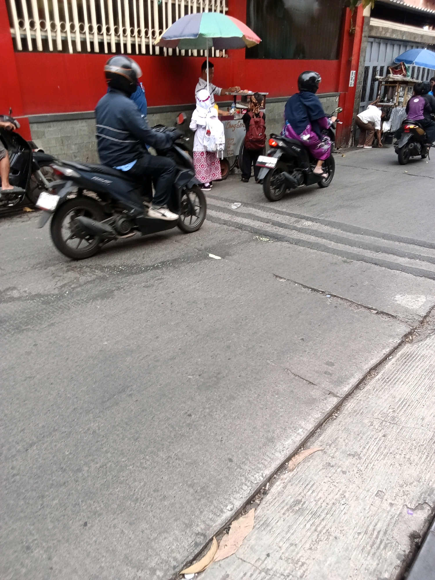 The image depicts a street scene, likely in Bandung City, Indonesia, based on the presence of a street vendor with an umbrella and a cart. In the foreground, a person wearing a black helmet and a blue jacket is riding a black motorcycle. The motorcycle has a license plate that is difficult to read. Behind this motorcycle, another person in a black helmet is riding a black motorcycle with a purple garment draped over them. Further in the background, a person in a white shirt and brown pants is bent over, seemingly interacting with a street cart. There are other individuals visible in the background, including someone wearing a purple top and a person with a red backpack. The environment appears to be a typical urban street with a red wall and a gate in the background. The road surface is asphalt, with some visible tire marks and debris. The overall impression is of a busy street with everyday activities taking place.