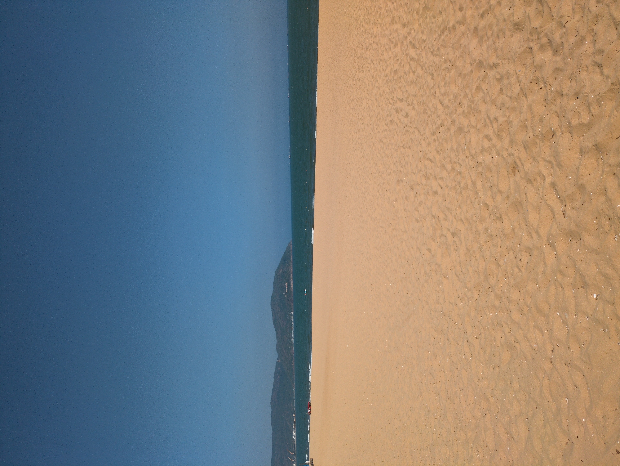 The image depicts a view from above of a sandy beach adjacent to the ocean in the city of Quy Nhon Ward, Vietnam. The horizon is a vertical line between the tan-colored beach and the dark blue sky. Between the sky and the sand is a line of water. A large, rocky hill rises along the shoreline of the water. The beach shows a pattern of footprints and scattered debris.