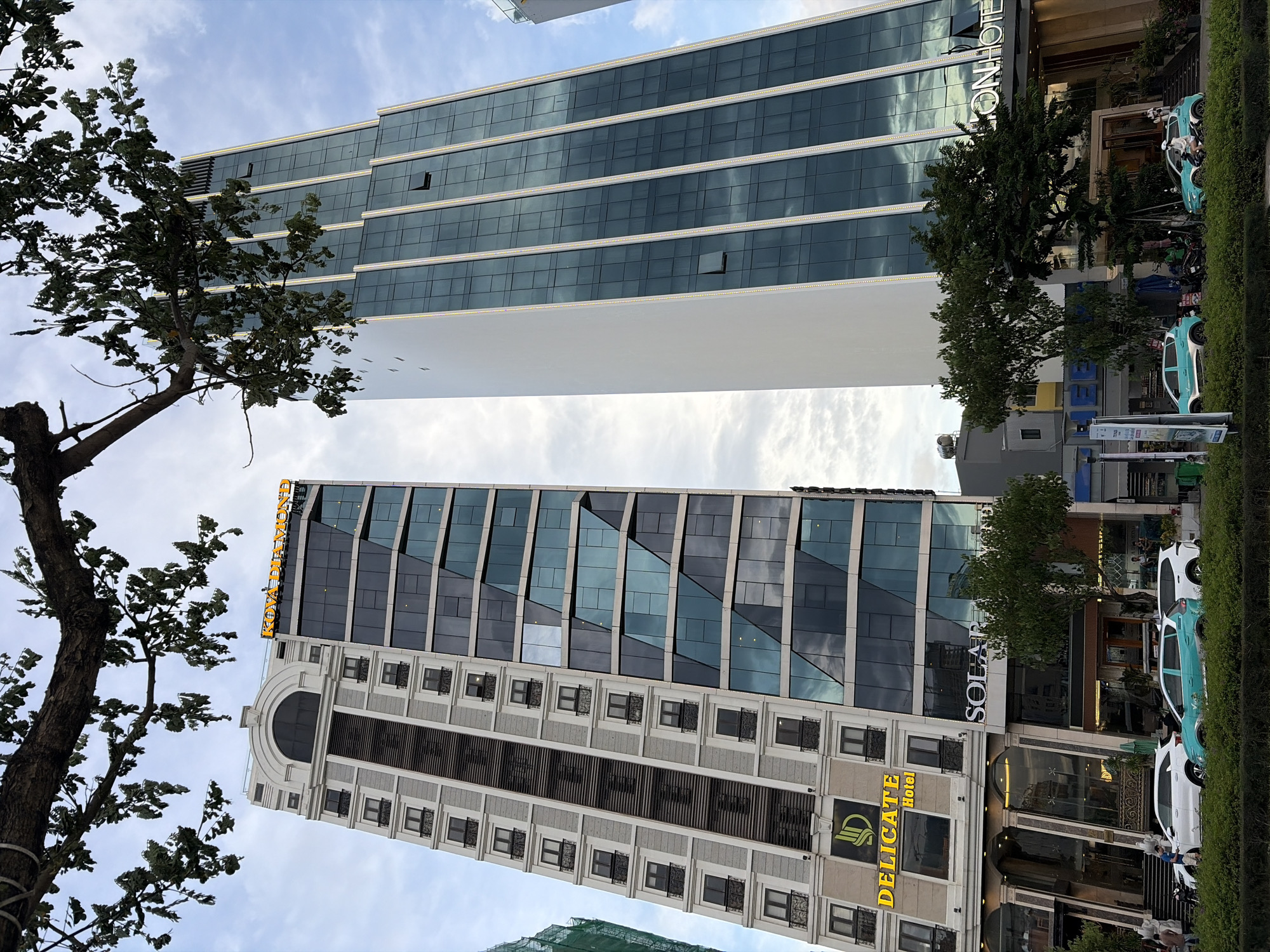 A daytime, low-angle, eye-level shot captures two modern skyscrapers in Da Nang, Vietnam, with a tree branch in the foreground. The skyscraper on the left is a mixed-use building with a white facade and a section of dark glass. The name "DELICATE Hotel" is prominently displayed in yellow on the white section. The skyscraper on the right is a taller, more slender building with a white side and a large section of dark, reflective glass, with "ON HOTEL" visible on its lower section. Several cars, including a white SUV, a teal hatchback, and a white hatchback, are parked in front of the buildings on the street. Pedestrians are visible on the sidewalk, and lush green bushes line the street. The sky is partly cloudy.