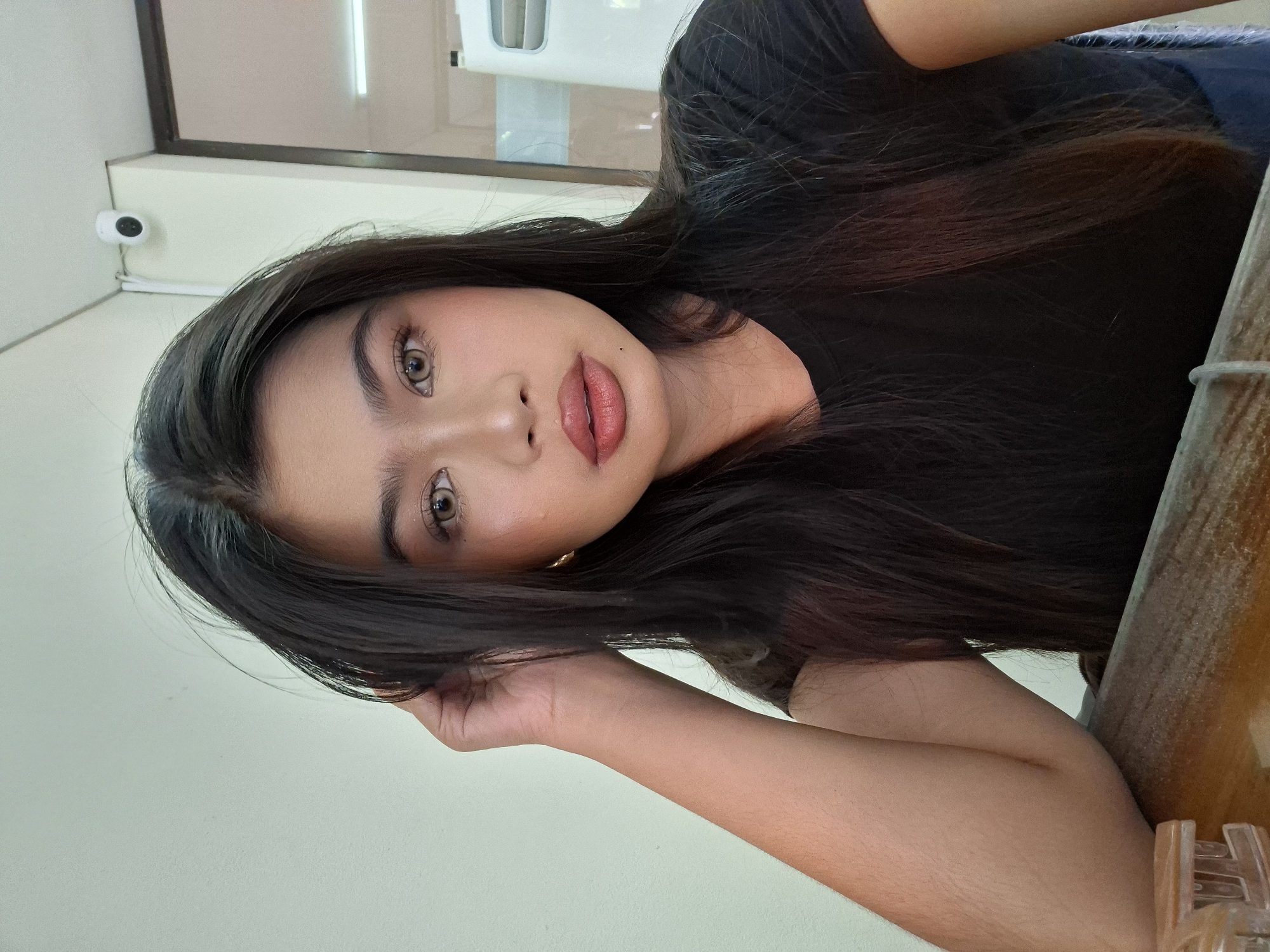 A young woman with long, dark hair is captured in a close-up selfie, leaning her head on her left hand. She is wearing a black t-shirt. Her makeup is visible, with emphasis on her eyes and lips. The background is a plain, light-colored wall, with a security camera visible in the upper right corner, and a glimpse of a door and a white air conditioner unit. A wooden surface is in the foreground, with a clear plastic object on the left. This scene is indoors, likely in a public or semi-public space within Kuta, Indonesia, such as a cafe, restaurant, or shop.