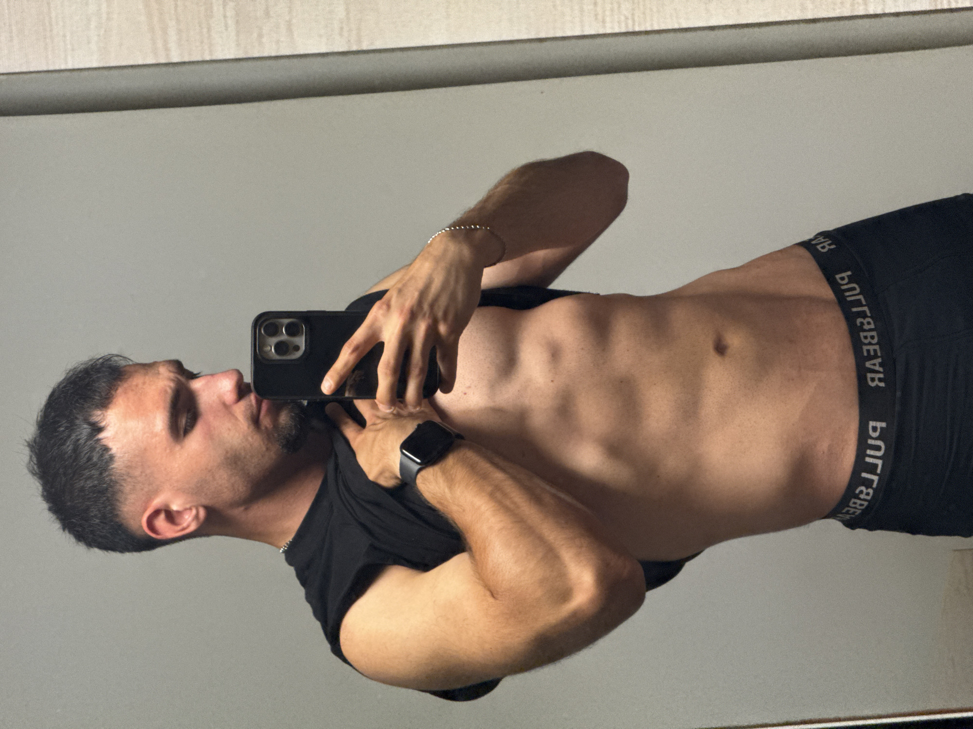 A young man is captured in a mirror selfie, presenting a head-to-waist view. He has short, dark hair and a trimmed beard, looking towards the smartphone he holds in his right hand. The phone is black with a distinctive multiple-camera array. He wears a black t-shirt pulled up to expose his toned abdomen and chest. Black boxer briefs are visible, with the mirrored and inverted text "PULL&BEAR" on the waistband. A black smartwatch is on his left wrist, and a silver beaded bracelet adorns his right. The background reflected in the mirror is a plain light gray wall, with a horizontal dark bar and a light wood-textured surface visible above it. The image is evenly lit, suggesting an indoor setting. The man's expression is neutral, focused on the camera. The scene suggests a personal moment captured in a private space within Shumen, Bulgaria.
