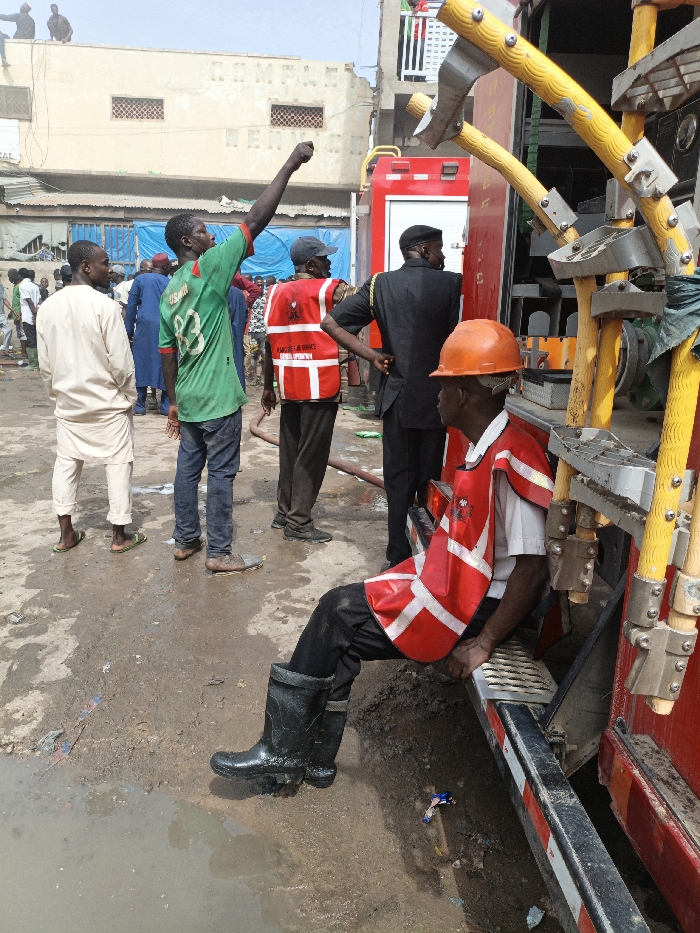 The scene depicts an outdoor urban environment in Fagge C, Nigeria, during daylight hours. A red fire truck is positioned on the right side of the frame.
In the immediate foreground, a male individual is seated on a step of the fire truck, facing right, partially out of view. He wears an orange hard hat, a white shirt, a red safety vest with prominent white reflective stripes, black trousers, and black rubber boots. His left leg, clad in a rubber boot, extends towards a muddy puddle on the ground.
Several other individuals are present in the midground and background. A male subject, central to the midground, wears a green short-sleeved shirt with "USMAN 83" printed on the back and blue jeans. He has his right arm raised with a clenched fist, gesturing upwards and to the left. To his left, another male wears a light-colored, long-sleeved traditional tunic and trousers with green flip-flops.
Behind the gesturing man, two emergency personnel are visible. One wears a red safety vest with "KANO FIRE SERVICE GENERAL OPERATION" text, dark trousers, and a dark cap, standing with hands on hips. Adjacent to him, another male in a dark suit and dark cap stands with his back largely to the camera. A dark hose is visible on the wet, muddy ground near these personnel.
The ground surface is uneven, consisting of mud and standing water puddles, with scattered litter including plastic wrappers. In the background, a light-colored, multi-story building with small rectangular windows is visible. Faint lettering, possibly "COMPANY LTD," is discernible on its facade. Two individuals are situated on the rooftop of this building. Another building with red and white structural elements is partially visible behind the fire truck.
