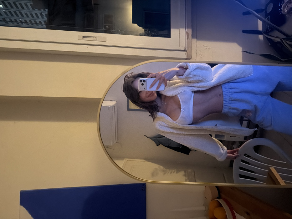 A young female subject is depicted taking a selfie in a large, arch-shaped mirror with a gold frame. The scene is set indoors, within what appears to be a residential space located in the city of Tel-Aviv, Israel. The subject, possessing medium-length dark hair, is smiling and holds a white smartphone in her right hand, positioned to photograph her reflection. She is dressed in a white bralette, a cream-colored fuzzy zip-up jacket, and light blue wide-leg pants. A delicate necklace is visible around her neck.
The interior environment features light-colored walls. To the left, a large rectangular blue abstract artwork with a small marking ("669") is affixed to the wall. Reflected in the mirror above the subject's head is a wall-mounted air conditioning unit. Further left in the reflection, a white slatted chair is visible, along with a wooden surface holding a bowl of oranges and another container. Behind the subject's reflection, a dark jacket hangs. To the right of the mirror, a narrow window reveals an urban night scene with multiple illuminated floors of tall buildings. The room is illuminated by warm, artificial light, and a prominent blue light source emanates from the lower right side of the frame.