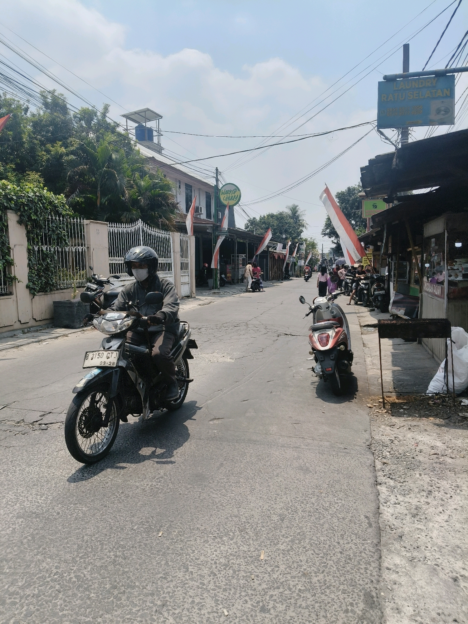 The image depicts a street scene in South Tangerang, Indonesia, on a sunny day with scattered clouds. The street is paved with asphalt and shows signs of wear and tear with cracks and uneven surfaces. On the left side of the street, there is a white fence covered in greenery, and beyond it, a building with a roof-mounted water tank. Several Indonesian flags, red and white, are visible along the street, suggesting a patriotic or celebratory occasion.
In the foreground, a person wearing a dark jacket, helmet, and face mask rides a black motorcycle. The motorcycle has a license plate that reads "B 3150 GTJ" and the registration date "09-28". The rider's hands are gloved.
Further down the street, several other motorcycles are parked or in motion. On the right side of the street, there are small businesses or stalls with awnings, and some people are gathered near them. One stall has a sign that says "MASAKAN PADANG" (Padang cuisine). A blue sign for "LAUNDRY RATUSELATAN" is also visible, along with contact numbers. A red and white scooter is parked on the right side of the road. In the background, more buildings and trees are visible. The overall impression is of a typical bustling street in an Indonesian city.