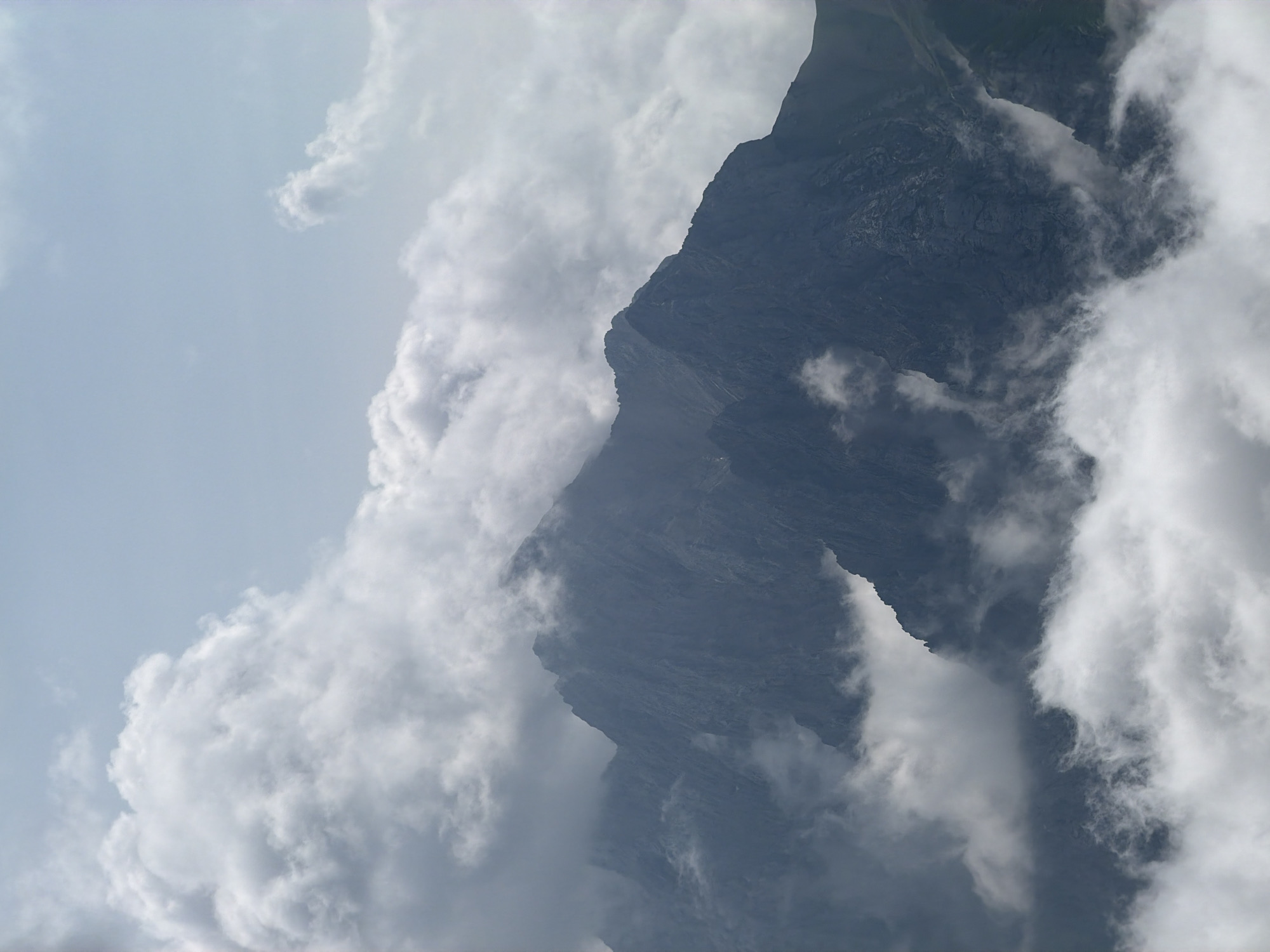 This is a close-up, slightly overcast, aerial photograph of the Zugspitze mountain in Garmisch-Partenkirchen, Germany. The image captures the jagged, dark grey, rocky peak of the mountain partially obscured by soft, white, billowing clouds. The clouds are dense and appear to be surrounding the upper portions of the mountain, creating a dramatic and somewhat mysterious atmosphere. The sky above is a pale blue, with lighter wisps of clouds scattered throughout. The terrain appears rugged and mountainous, with visible textures of rock formations. There are no people or man-made objects visible in the image.