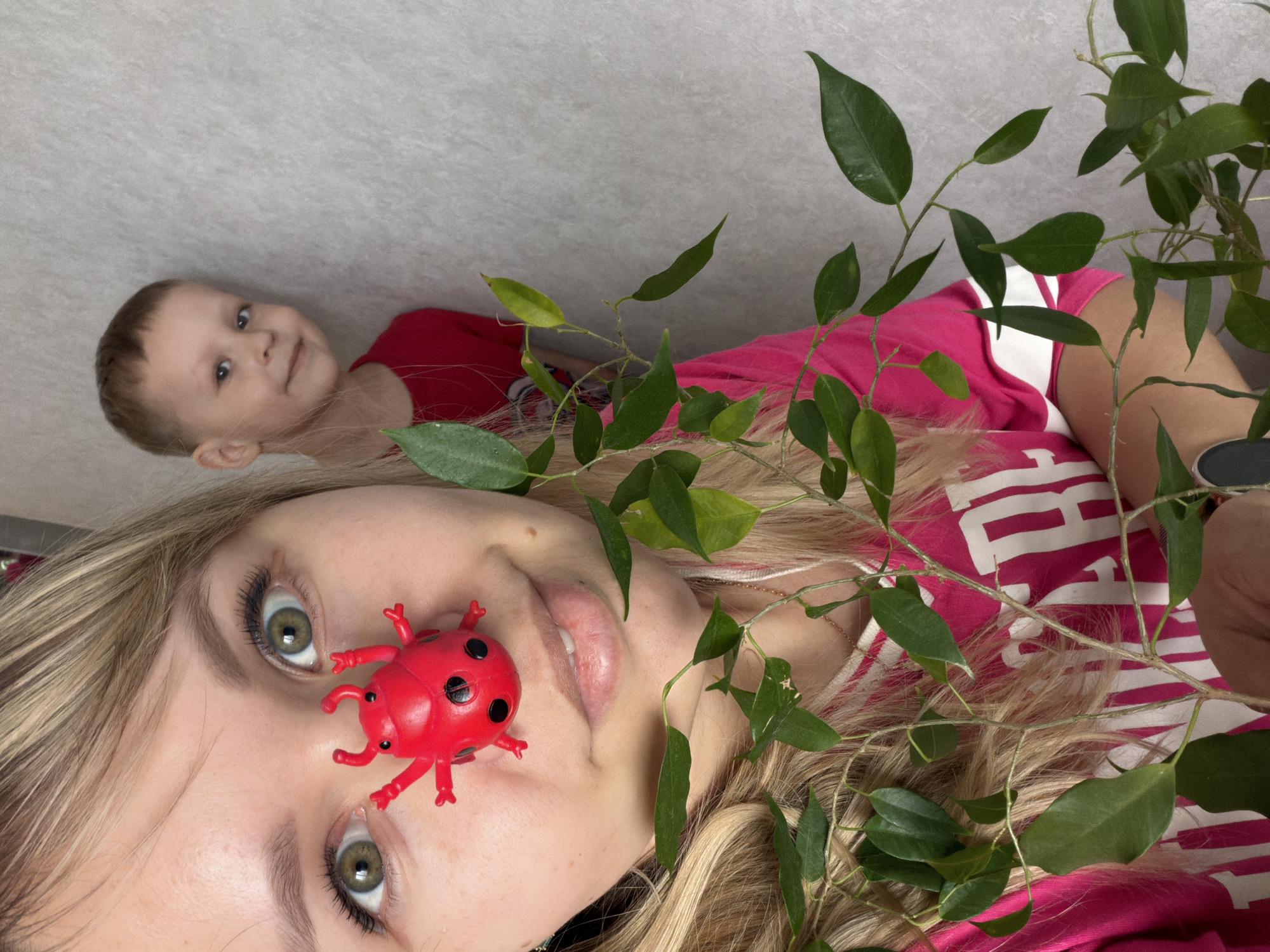 The image shows a woman lying down with a red toy ladybug on her nose. She is wearing a pink and white striped shirt, and there is a green plant with leaves around her. In the background, a young child is also lying down, wearing a red shirt. The background appears to be a light gray wall.