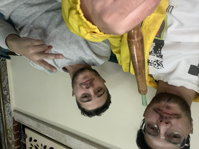 This image depicts two young adult males in an indoor setting within the city of Homs, Syria. The male in the left foreground, who appears to be taking the photograph, wears round clear-framed eyeglasses, a white long-sleeved shirt featuring a black and white graphic print, and a bright yellow jacket. He has dark hair, a mustache, and a beard, and his gaze is directed towards the camera. He holds a wooden hookah hose mouthpiece with a green tip in his left hand. To his right, the second male is seated, wearing a grey long-sleeved top. He has short dark hair and a beard, and he smiles at the camera while making a 'V' gesture with his right hand. The background consists of light-colored walls, with decorative elements visible on the right, including a panel with intricate patterns, possibly latticework, and a portion of patterned wallpaper. A dark green or teal colored cushion or upholstery is partially visible behind the second male.