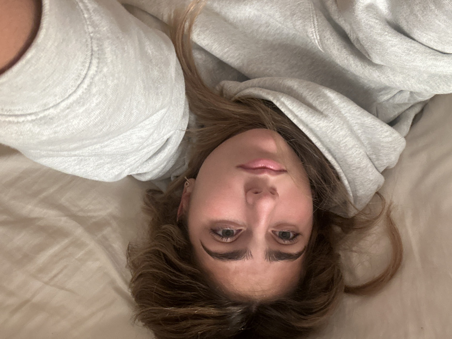 A young adult female is depicted lying supine on a light-colored, rumpled surface, likely bedding, in an indoor environment. The precise location within Aldoar, Foz do Douro e Nevogilde, Portugal, cannot be visually determined from the image. She is positioned diagonally across the frame, with her head in the lower-left quadrant and her torso extending towards the upper-right. Her head is slightly tilted, and she is looking directly towards the viewer. She has medium brown hair, fair skin with a slight flush on her cheeks, light-colored eyes, and prominent dark eyebrows. A small, thin hoop earring is visible in her left ear. She is wearing a light grey hooded sweatshirt. The lighting is soft and even, originating from an unidentifiable source. No other subjects or objects are visible in the plain background. No overt actions or interactions are occurring beyond the individual resting.