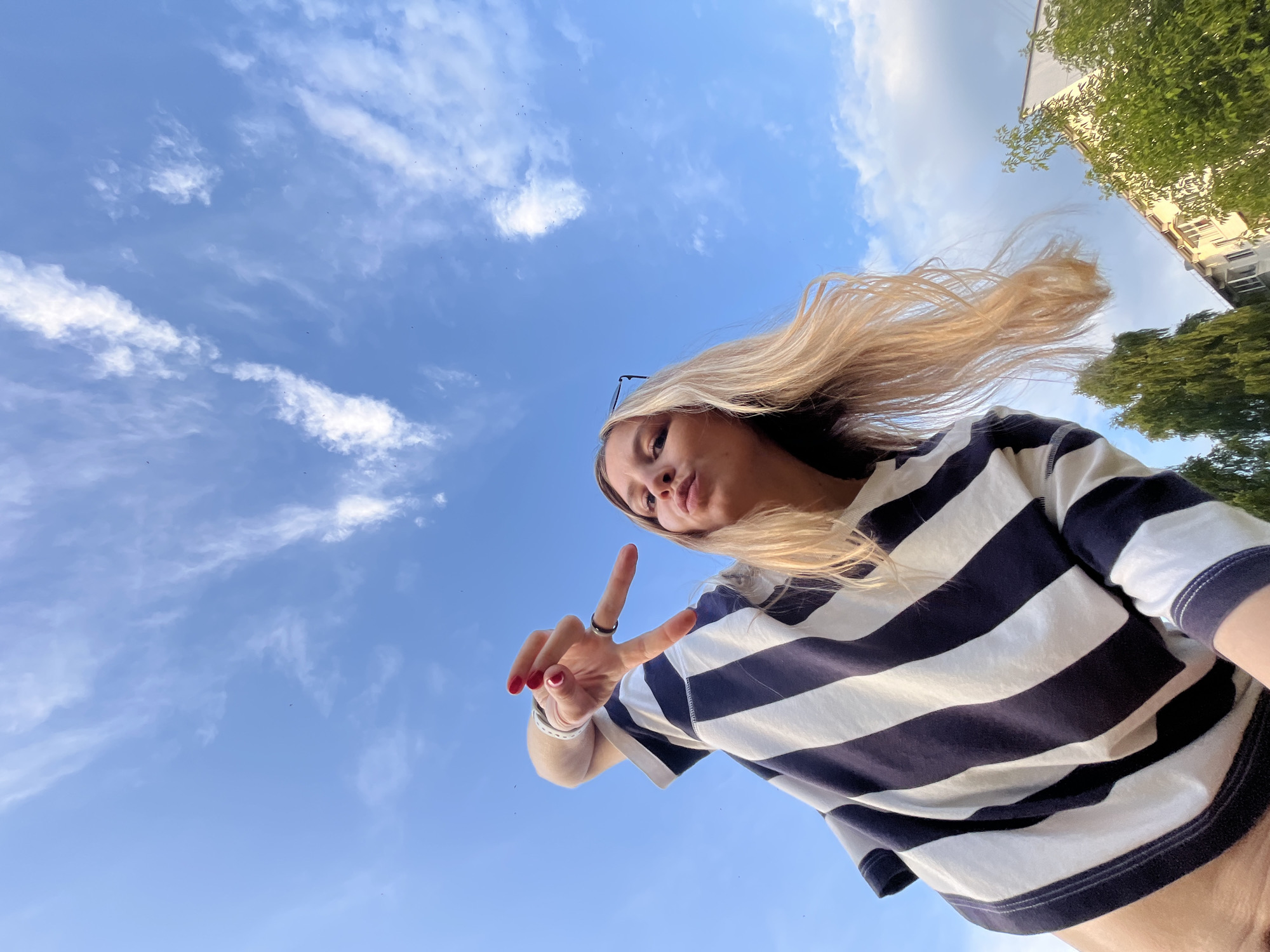 A young woman with blonde hair and red nail polish is posing for a picture outdoors. She is wearing a navy and white striped long-sleeved shirt and making a peace sign with her right hand. Her face is partially obscured as she pouts her lips and looks towards the camera. The background is a blue sky with scattered white clouds. In the lower right corner of the image, the tops of green trees and a building are visible. This image was taken in the city of Chaikovsky, Russia.