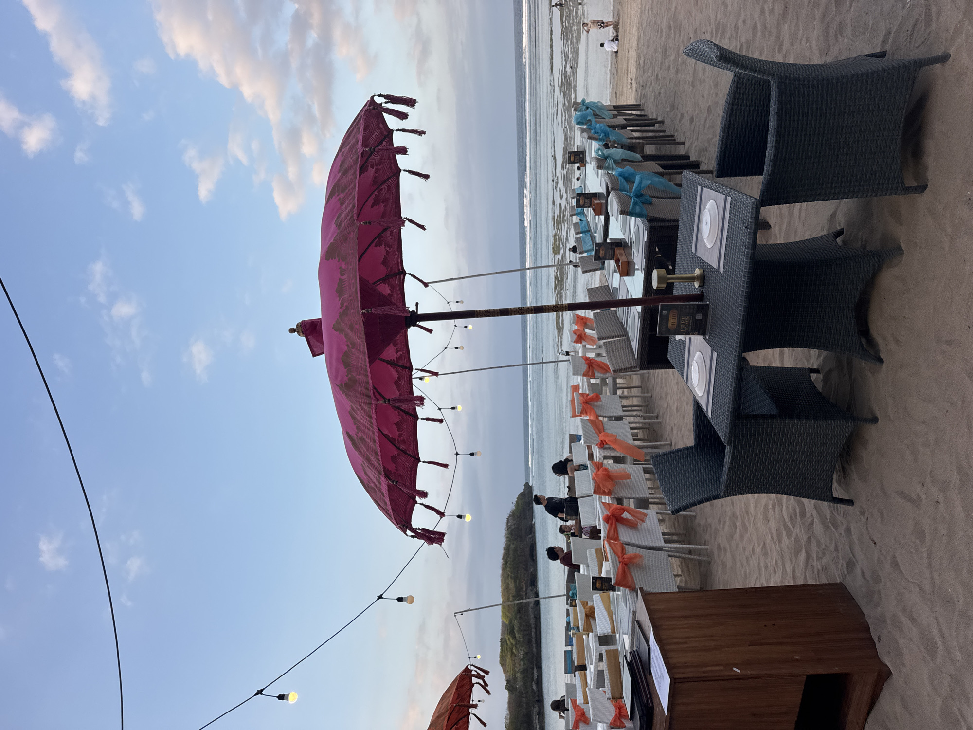 An outdoor dining area is set up on a sandy beach in Kuta Selatan, Indonesia, near the ocean. The scene is illuminated by string lights hanging overhead. Several tables are covered with white tablecloths and set with place settings. The chairs around the tables are white with orange and blue ribbons tied to them.
In the foreground, a dark woven table and chairs are positioned on the sand. A wooden cabinet with open shelves is to the left. Two large, ornate pink umbrellas provide shade over some of the tables. Another red umbrella is visible in the distance.
In the middle ground, several groups of people are seated at the tables, appearing to be dining or socializing. Others are walking along the beach. In the background, the ocean stretches out to the horizon, with gentle waves breaking on the shore. A small, rocky island or peninsula is visible in the distance on the left. The sky is a pale blue with scattered clouds.