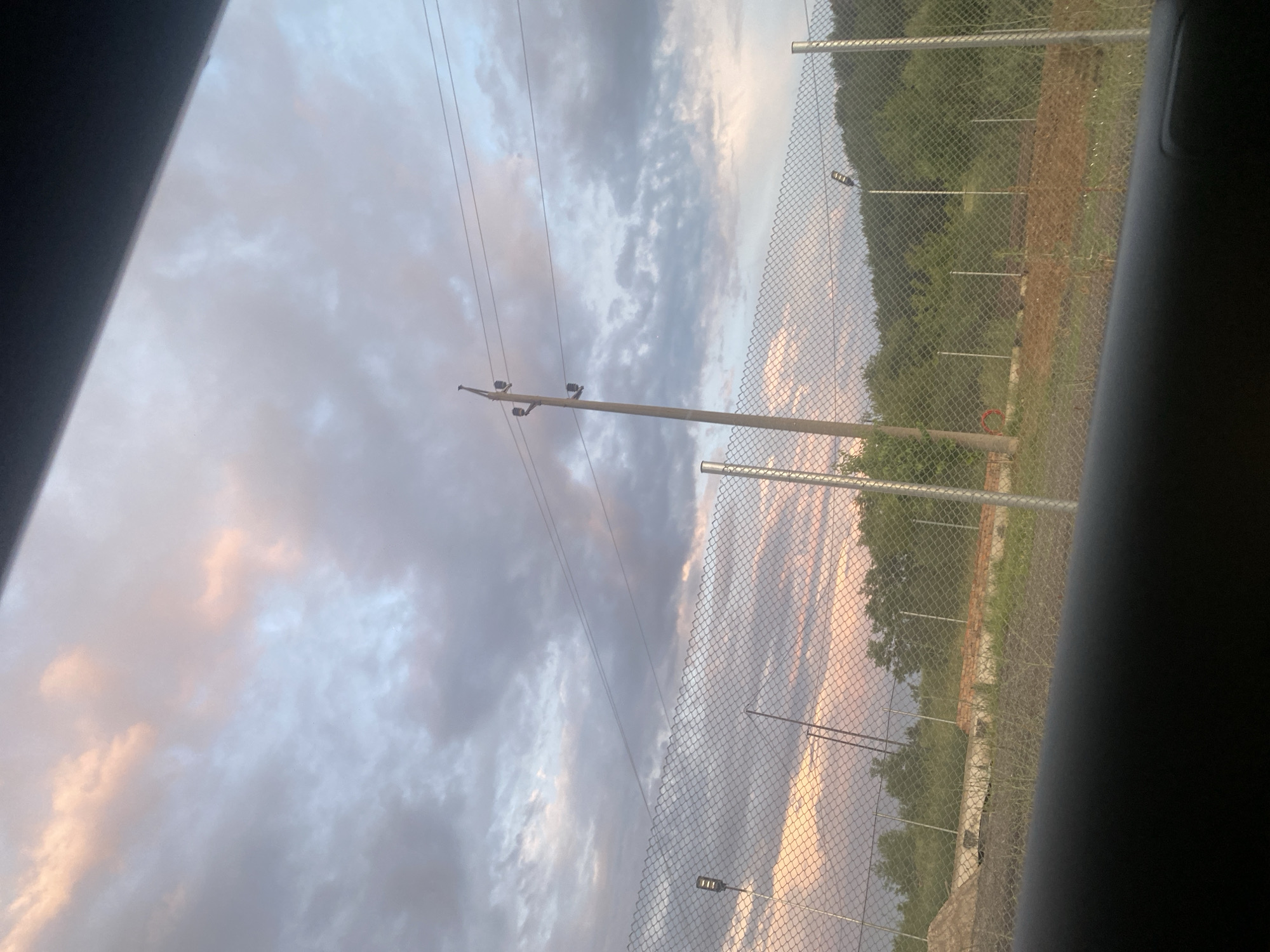 The image depicts a cloudy sky with patches of pink and pale blue hues, suggesting either sunrise or sunset. In the foreground, a chain-link fence stretches horizontally across the frame. Behind the fence, a landscape unfolds with trees and hills in the distance. A wooden utility pole with power lines is visible on the right side of the frame. There are also what appear to be streetlights or security lights mounted on poles scattered throughout the scene. The ground in front of the fence seems to be unpaved, with some areas of dirt and patchy grass. The overall impression is an outdoor scene, possibly on the outskirts of a town, given the combination of utility infrastructure and natural landscape. No people are visible in the image. The vantage point appears to be from inside a vehicle, indicated by the dark, blurry foreground element.