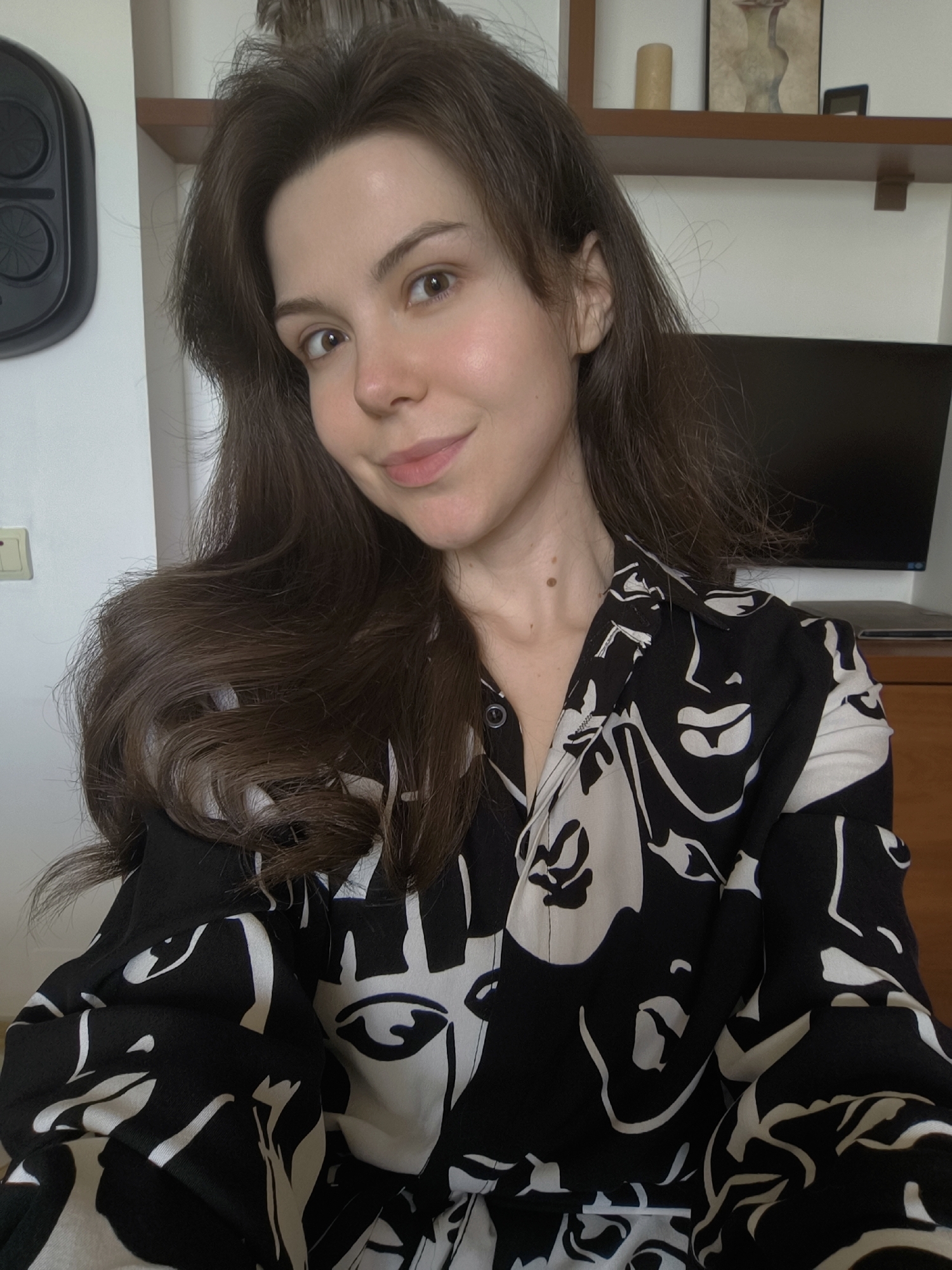 A young woman with long, dark brown hair and fair skin is pictured in a close-up, waist-up shot. She is looking directly at the camera with a gentle smile. Her eyes are brown, and her lips are slightly parted. She is wearing a black and white patterned shirt with an abstract face design. The background is blurred but suggests an indoor setting, with shelves and a television visible. There is no specific indication of a city or location.