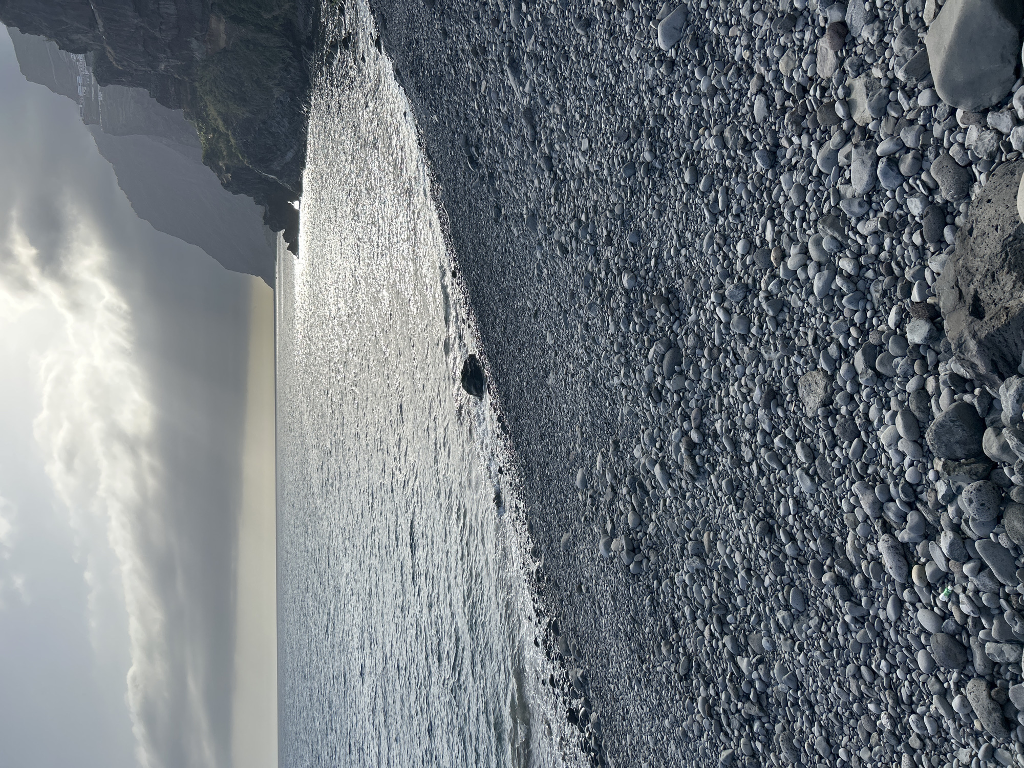 The image depicts a rocky beach in Câmara de Lobos, Portugal. The right side of the frame is dominated by a dense field of grey, rounded pebbles. The left side shows the ocean, reflecting the sunlight and appearing silvery. A cliff face is visible in the top left corner, disappearing into a cloudy sky.