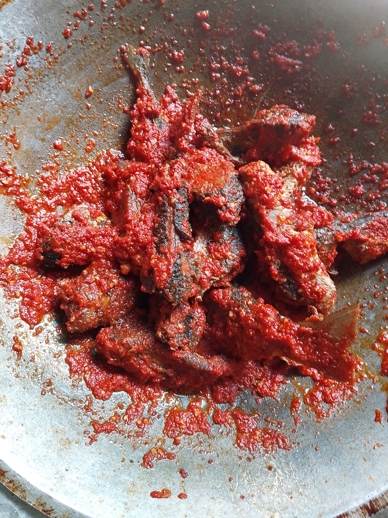 A close-up overhead shot captures small, fried fish generously coated in a vibrant red chili sauce, commonly known as sambal, within a metal wok or frying pan. The pan's interior surface is visible around the food, showing a dull, metallic sheen with scattered specks and streaks of the red sauce and cooking oil.
The fish pieces appear to be dark brown and crispy beneath the thick, chunky sauce. The red chili sauce is bright and textured, suggesting it's freshly made from ground red chilies, possibly mixed with other spices like garlic or shallots. The food is evenly distributed, indicating it has been thoroughly tossed or stirred in the pan.
The scene depicts a cooked dish, likely a common Indonesian meal, possibly just finished cooking or being prepared for serving. The setting is inferred to be a kitchen in Lhokseumawe, Indonesia. No people, visible text, specific time of day, or weather conditions are discernible from the image.