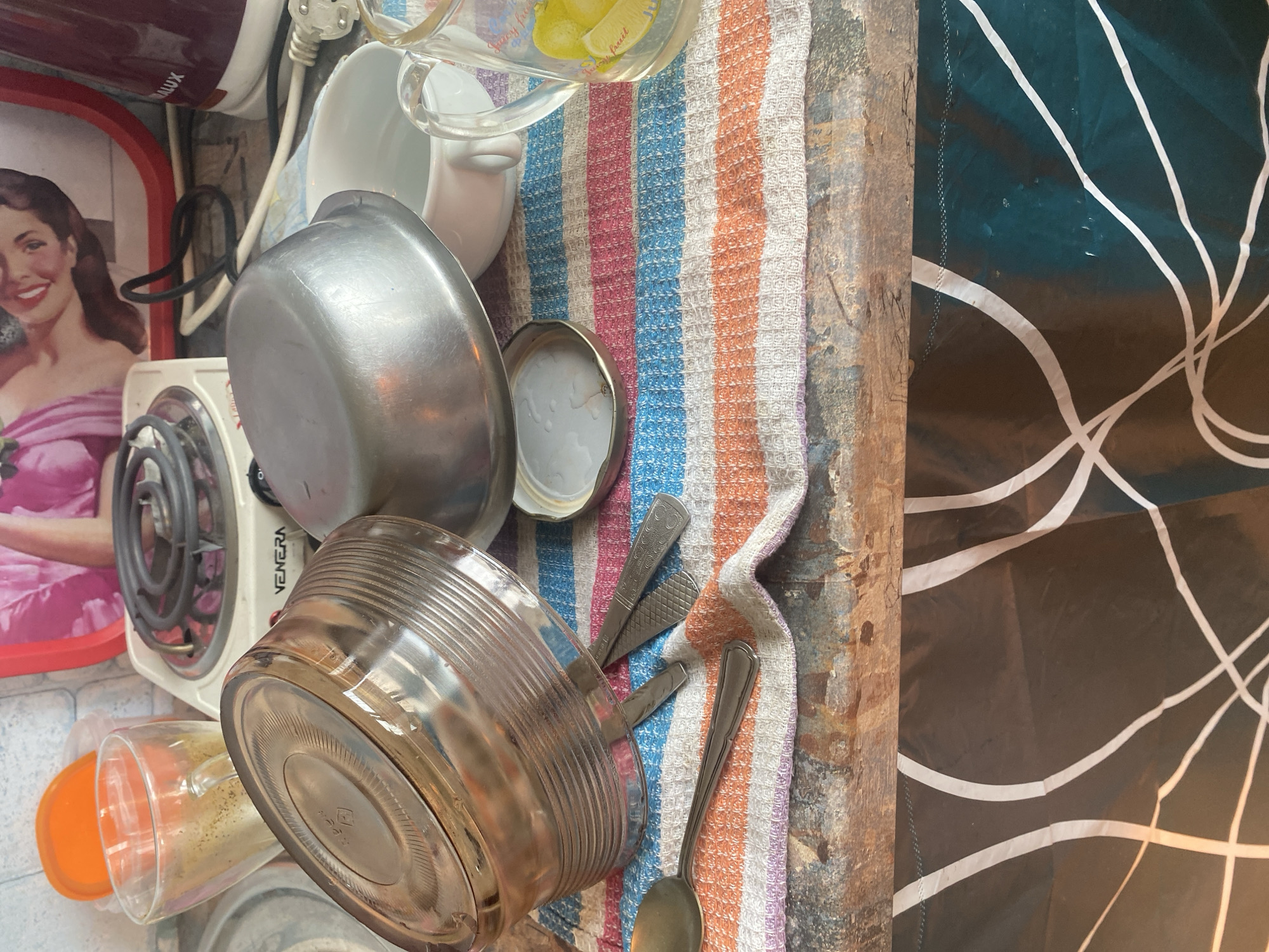 The image depicts a cluttered kitchen counter in Krupki, Belarus. In the foreground, a striped towel lays on a rustic wooden surface. On the towel, a large metallic bowl is placed upside down, revealing its ridged underside. Next to it lies a smaller metallic pot. A silver spoon and two small knives are also visible on the towel. Further back, a white ceramic mug and a glass pitcher with a lemon illustration are present. Behind these items, a small electric stove with a coiled heating element is partially visible, alongside a glass and an orange lid. A red tray with a picture of a woman is in the background. The overall scene suggests a domestic setting with items typically found in a kitchen.
