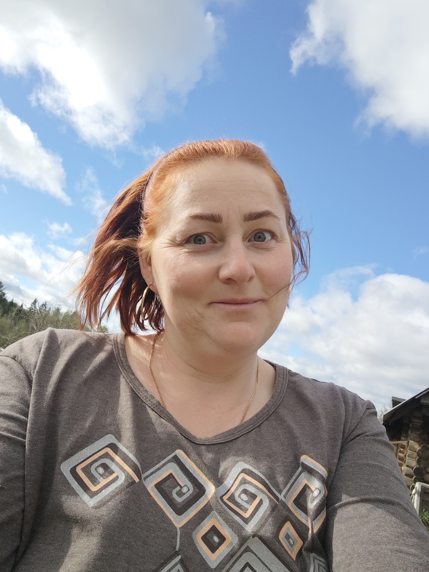 A low-angle, medium shot captures a woman with red hair pulled back in a ponytail, smiling slightly at the camera. She has fair skin and is wearing a gray t-shirt with a geometric design. A thin gold necklace is visible around her neck, along with hoop earrings. The background consists of a blue sky with scattered clouds. In the bottom right corner, a portion of a log cabin is visible. The photo appears to have been taken outdoors, possibly in Фоки, Russia, though the exact location within the city cannot be determined from the image.