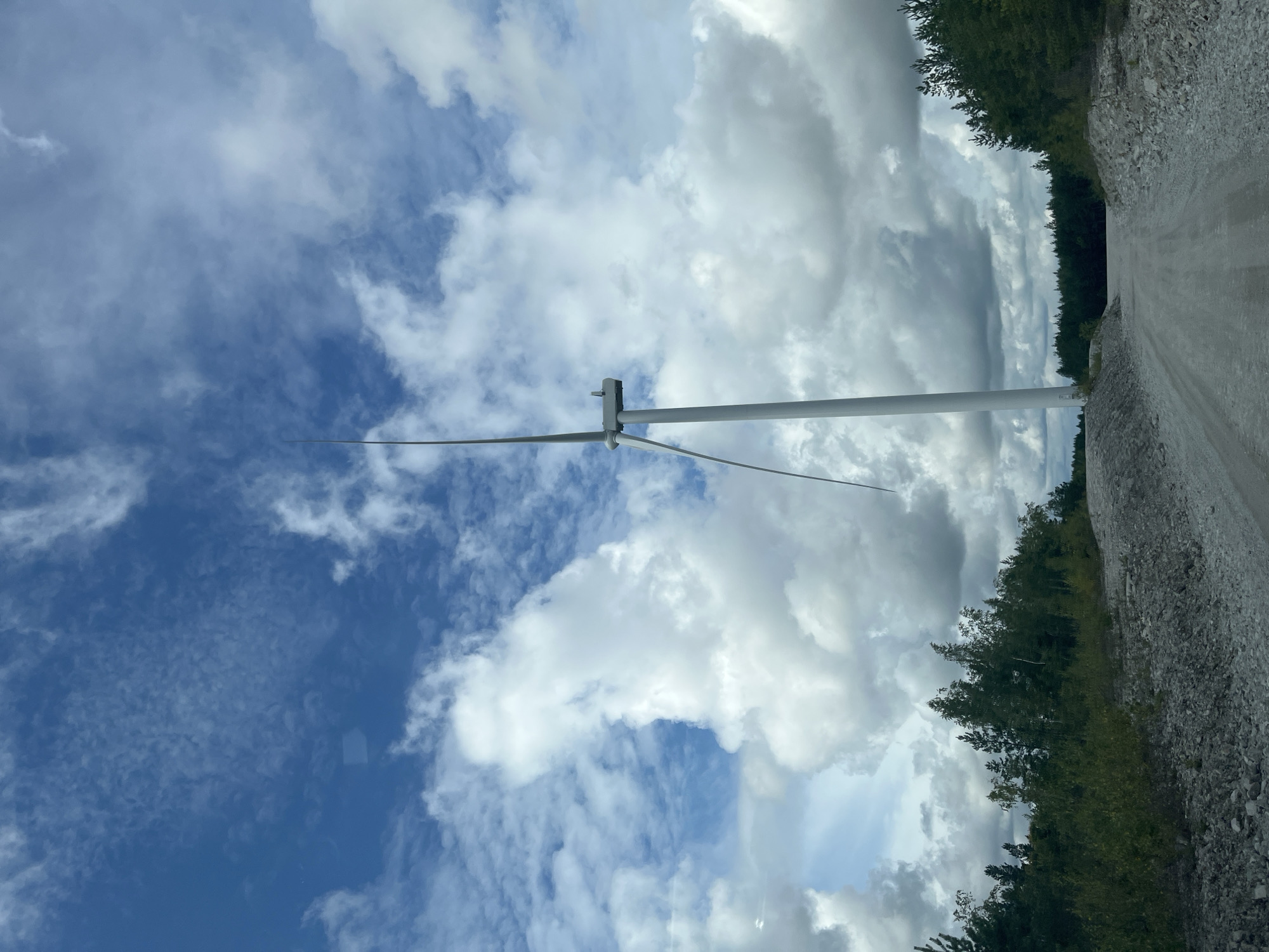 The image depicts a wind turbine in Aidu, Estonia. The wind turbine, a tall white structure with three blades, stands prominently against a bright blue sky filled with fluffy white clouds. The turbine is situated in a rural or industrial area, with a dirt road leading towards it. Dense green trees line the sides of the road and extend into the background. The ground in the foreground is covered with gravel and rocks, suggesting it might be a construction site or a service road for the wind turbine. The overall scene conveys a sense of open space and renewable energy infrastructure.