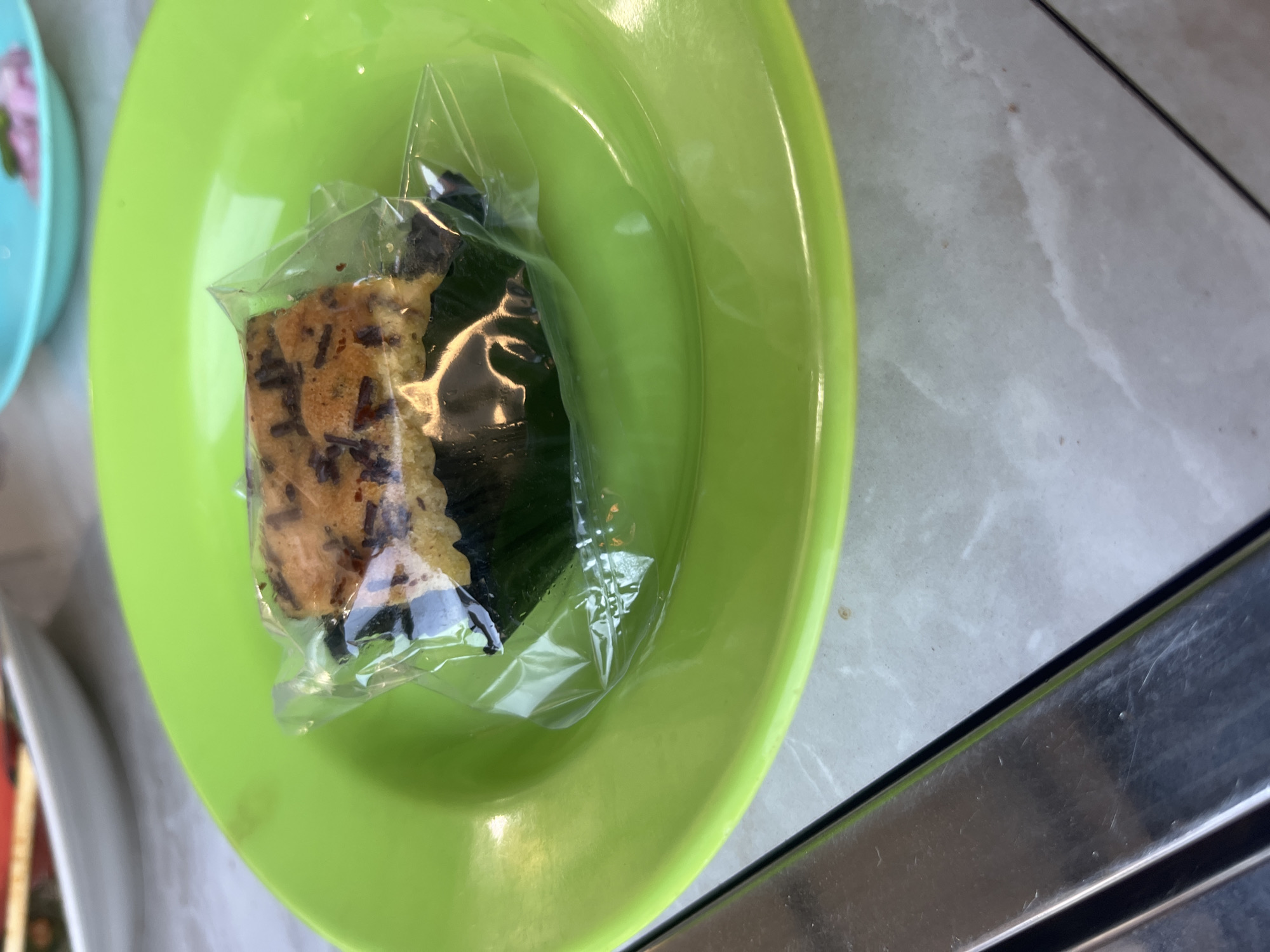A close-up, top-down view shows a slice of chocolate chip cake on a bright green plate. The cake is wrapped in clear plastic and rests in a dark green paper liner. The plate is positioned on a tiled surface. In the background, out of focus, are glimpses of other plates and tableware, including a teal plate with pink food and a white plate with red trim. A metallic object, possibly a table leg, runs along the bottom left of the frame. The scene appears to be indoors, possibly in a restaurant or cafe setting in Glameunasah Baro, Indonesia.