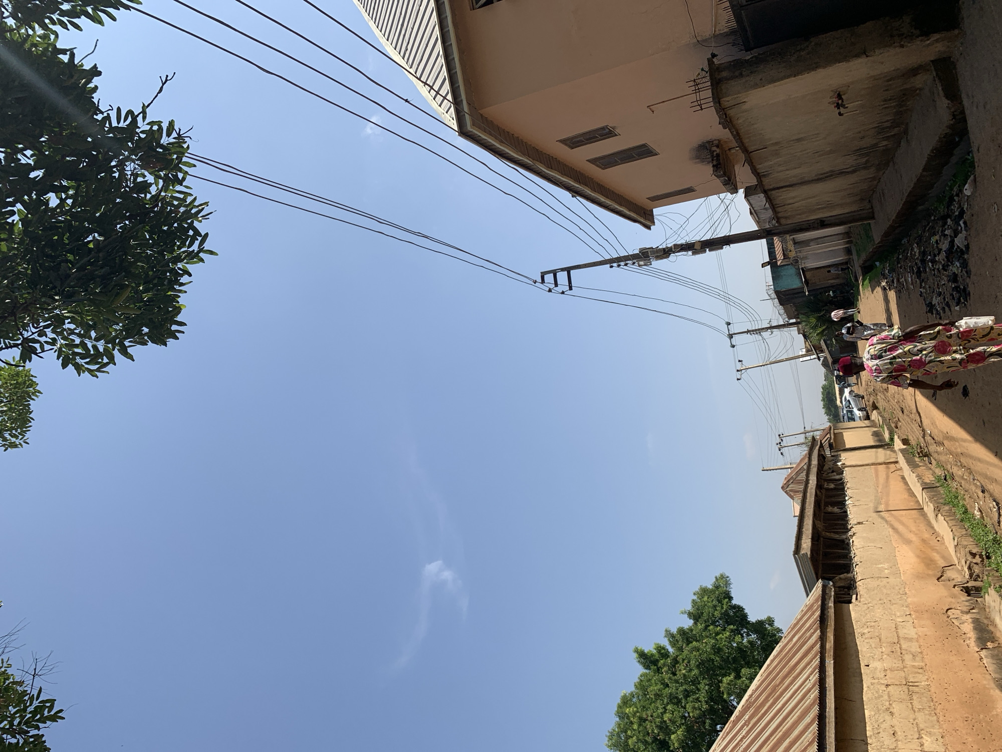 The image shows a street in Rafin Par, Nigeria, with a blue sky above. Power lines run across the sky. Buildings line the street, with one being light brown and the other being a weathered concrete wall. People are walking down the street. Trees are visible on both sides of the street. There is some litter and debris along the side of the road.
