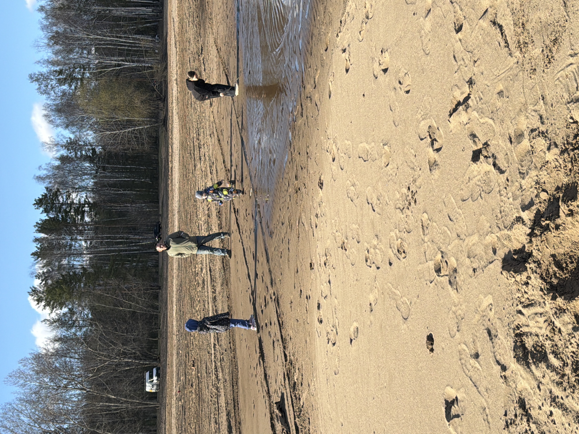The image depicts a sandy area next to a forest. Four people are present, all facing to the left. A person in black is standing in a puddle; another person is next to them. A third person is wearing green clothing and is looking to the right. A fourth person is wearing a blue hat. A white vehicle is parked on the edge of the forest in the distance. The ground is covered in footprints. This location is in the city of Chaikovsky, Russia.