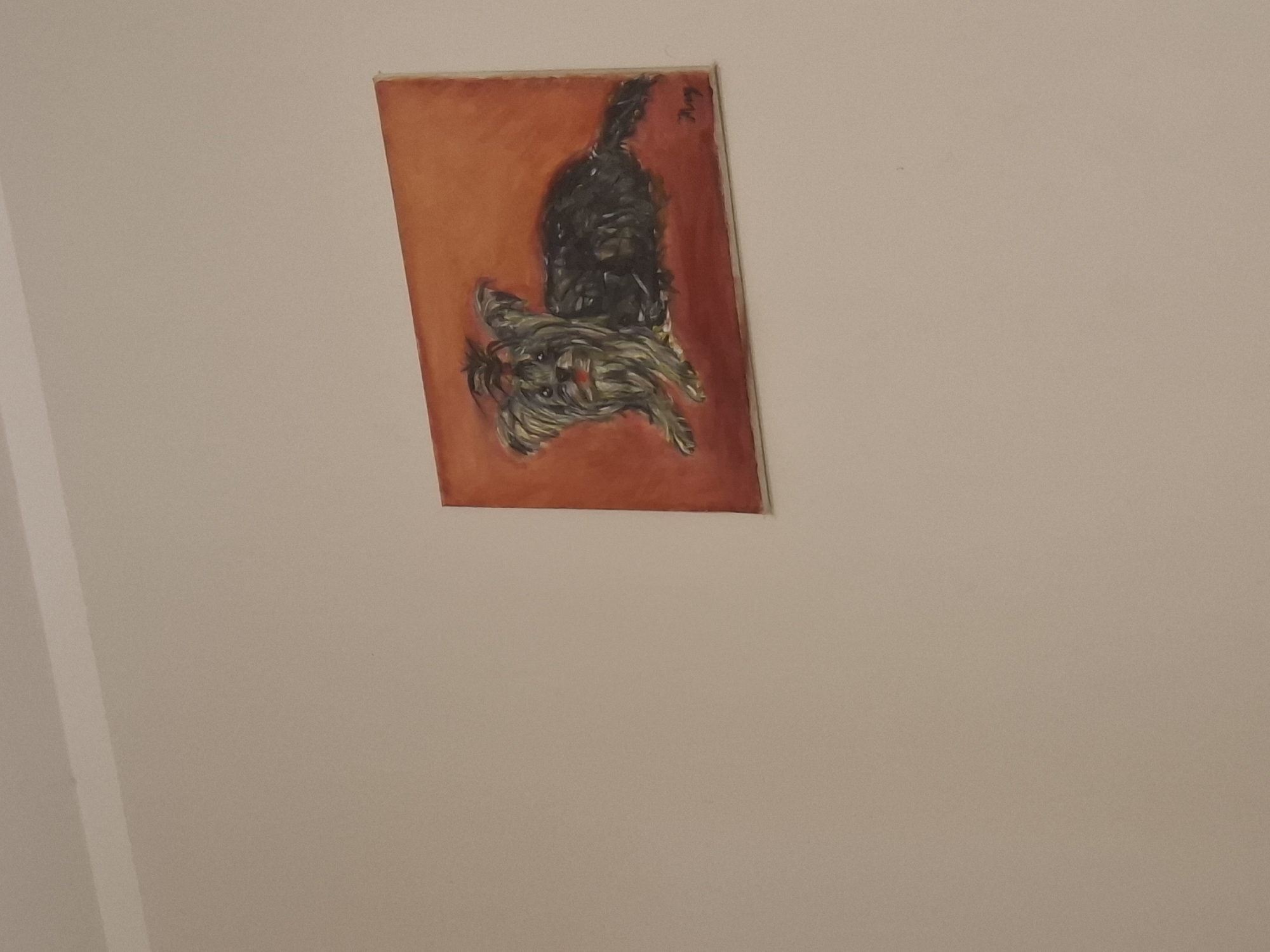 A slightly angled, medium-shot photograph shows a framed painting hanging on a pale wall. The painting, rectangular and vertically oriented, features two dogs on a bright orange background. The dog closest to the viewer is smaller and appears to be lying down, with visible details of its face and legs. A larger, darker dog is positioned above it, its tail visible at the top of the painting. A signature is present near the top right corner of the artwork. The rest of the wall is empty and uniformly colored. The location is not mentioned.
