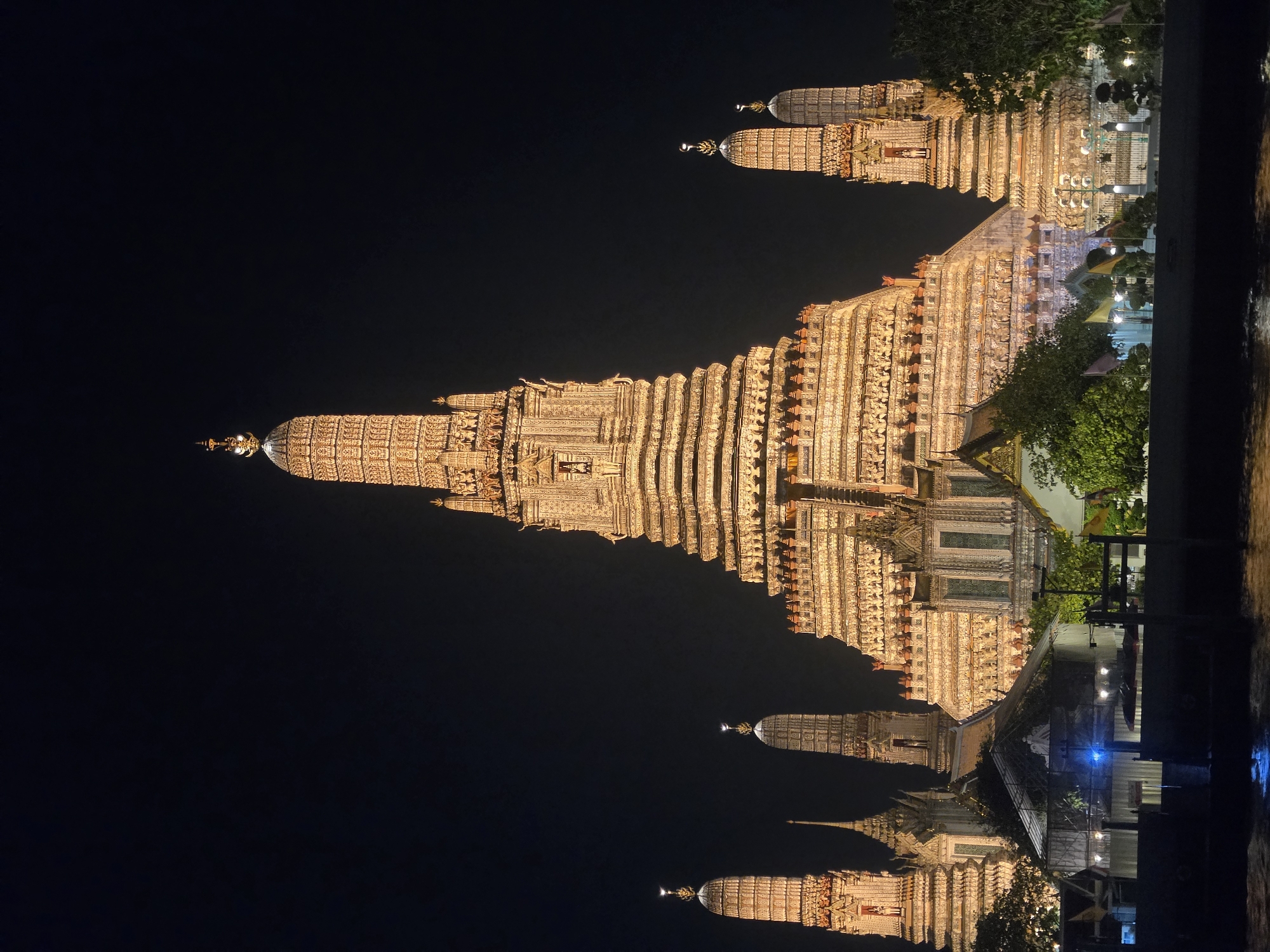 The image shows a night view of Wat Arun, a Buddhist temple in Bangkok, Thailand. The temple is illuminated, and its reflection is visible in the Chao Phraya River. The sky is dark.
