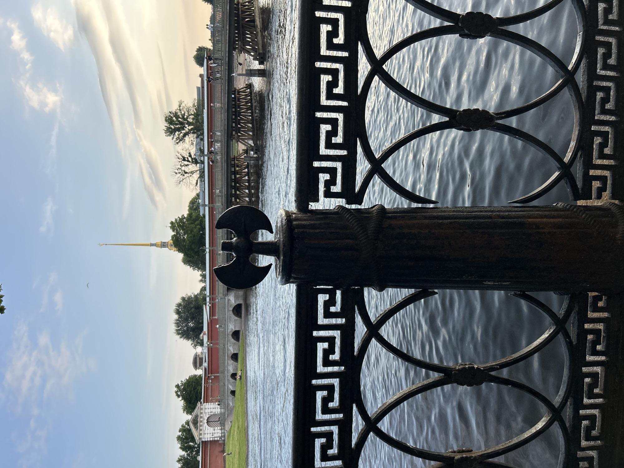 The image captures a view of a body of water with a decorative metal fence in the foreground. Behind the fence, across the water, stands a red brick fortress-like structure with arched openings, and a tall, golden spire piercing the sky. A wooden bridge spans the water, with people visible walking on it. Trees dot the landscape behind the fortress. The sky above is a pale blue with wispy clouds, suggesting it is either early morning or late afternoon. The water is rippling, reflecting the ambient light. The foreground fence features a repeating geometric pattern and is supported by a dark, ornate post topped with a decorative element resembling an axe. The overall scene suggests a historical or cultural landmark, possibly in a city with significant architectural heritage.