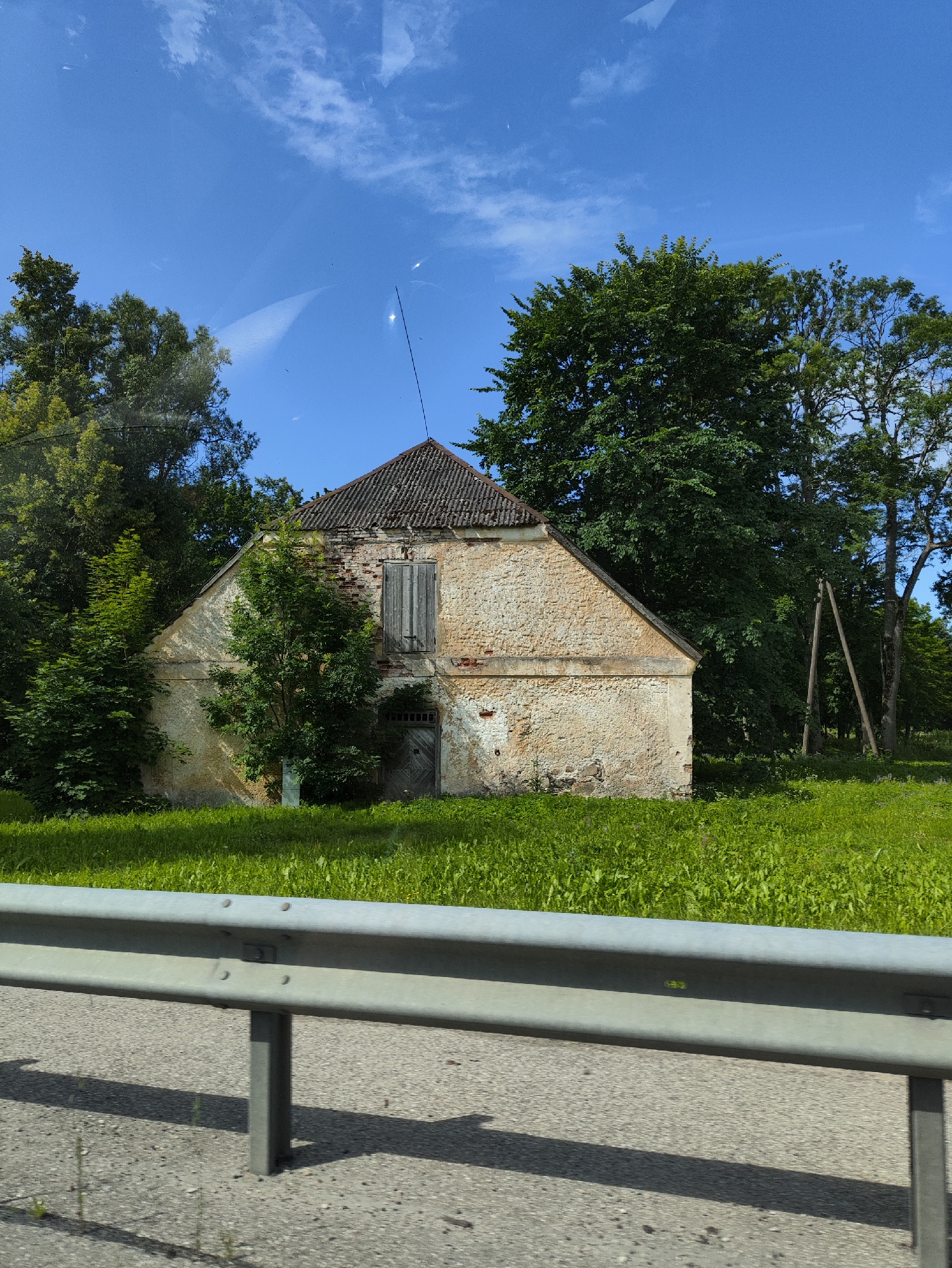 The image shows an old, abandoned building with a weathered stone facade and a decaying tiled roof, located in the rural area of Svētciems, Latvia. The building has wooden doors and shutters, and is surrounded by lush green grass and trees. In the foreground, a metal guardrail and asphalt road are visible, suggesting the building is situated near a road. The sky is a clear blue with a few scattered clouds, indicating a sunny day. There are no people or animals visible in the image. The overall impression is one of a neglected historical structure in a tranquil natural setting.