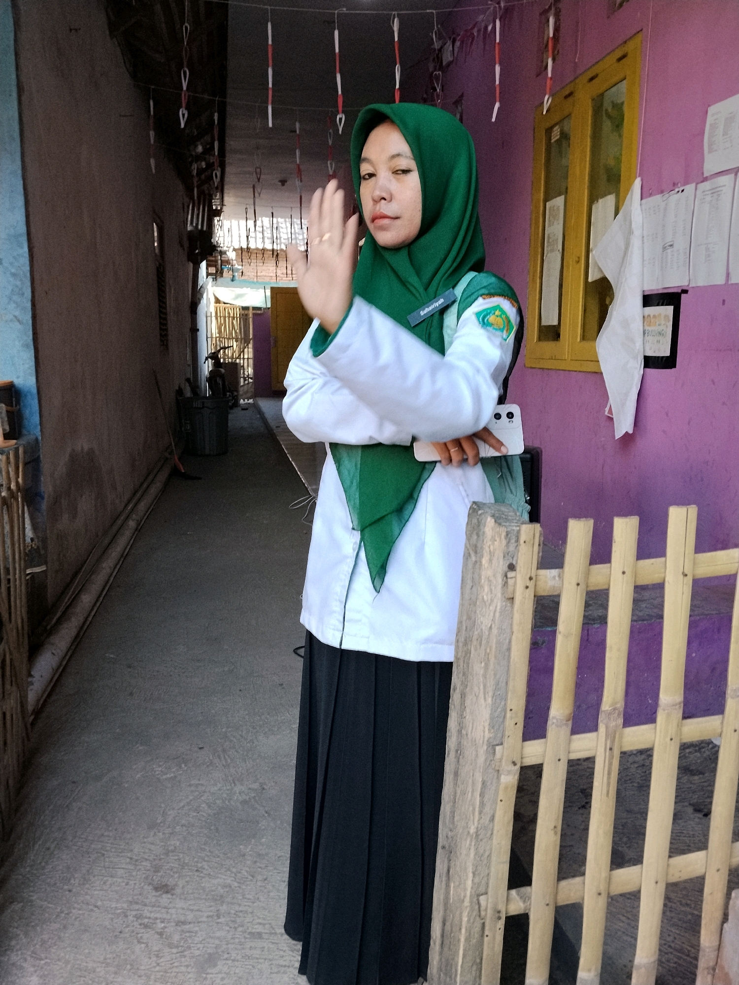 In the city of Panggungan, Indonesia, a young person wearing a green hijab and a white uniform with a green scarf stands in a narrow, open-air corridor. The corridor walls are a faded blue on the left and a bright pink on the right. A bamboo fence partially obscures the view on the right side of the frame. The person holds a white smartphone in their right hand and raises their left hand in a waving gesture. The ground is a concrete pathway. Hanging decorations, possibly festive, are strung across the ceiling of the corridor. In the background, a motorbike is visible next to a trash can. A yellow-framed window is present on the pink wall.