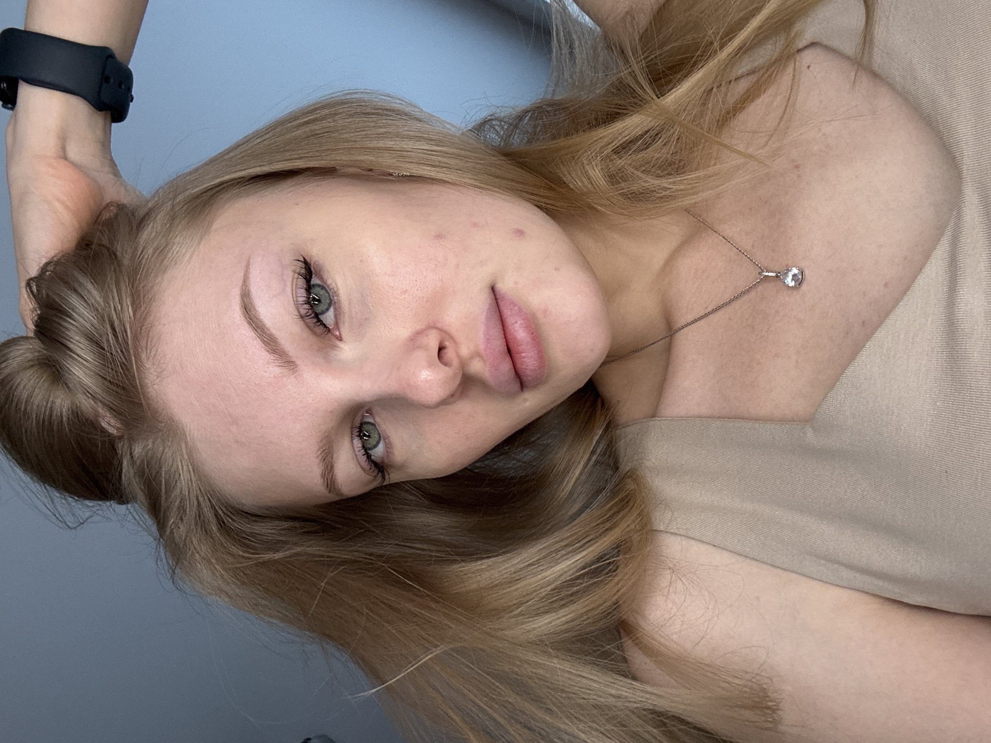 A fair-skinned, light-haired woman is pictured in a close-up, tilted portrait. She is wearing a beige top and a silver necklace with a clear pendant. A black smartwatch is visible on her wrist. The background appears to be a muted blue-grey wall. The location in Perm, Russia cannot be determined from this image.
