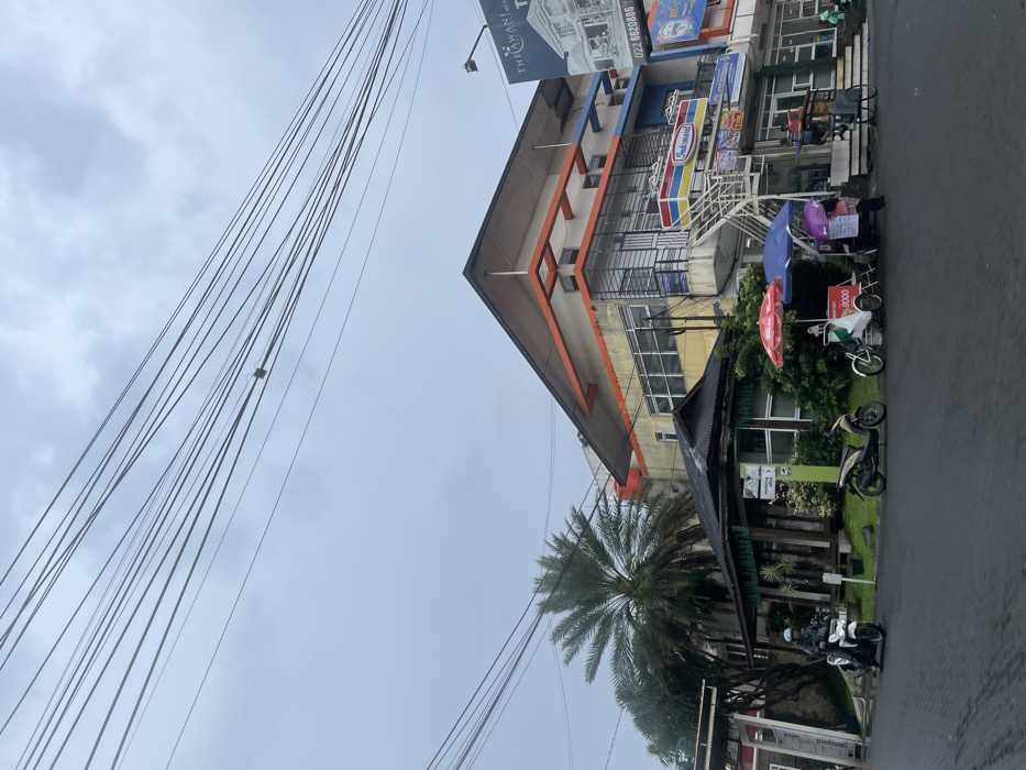 The scene is a street corner in Ngamprah, Indonesia (latitude: -6.863, longitude: 107.51). The street is paved and wet, likely from recent rain. The sky is overcast with a light grey color. Numerous power lines run across the sky. A tall palm tree stands on the left side of the street. Two motorcycles are parked on the street. In the foreground, two people are standing in front of a small shop with a red awning. Behind them is a small cart selling ice cream with a red and white canopy. A larger building with an orange roof is visible in the background. There is a small sign above the entrance to the building, but it is not visible in the image. The building has a yellow facade and large windows. There are several businesses with signage on the front of the building, including a sign advertising "Indomaret." There is a small sign that reads "022 6820886" which is possibly a phone number for the building. A white van is parked at the corner of the building. There are several people on the street. Some are walking, others are standing. The scene is busy with activity, but there are no signs of any violence or disorder. The overall mood of the scene is calm and peaceful.