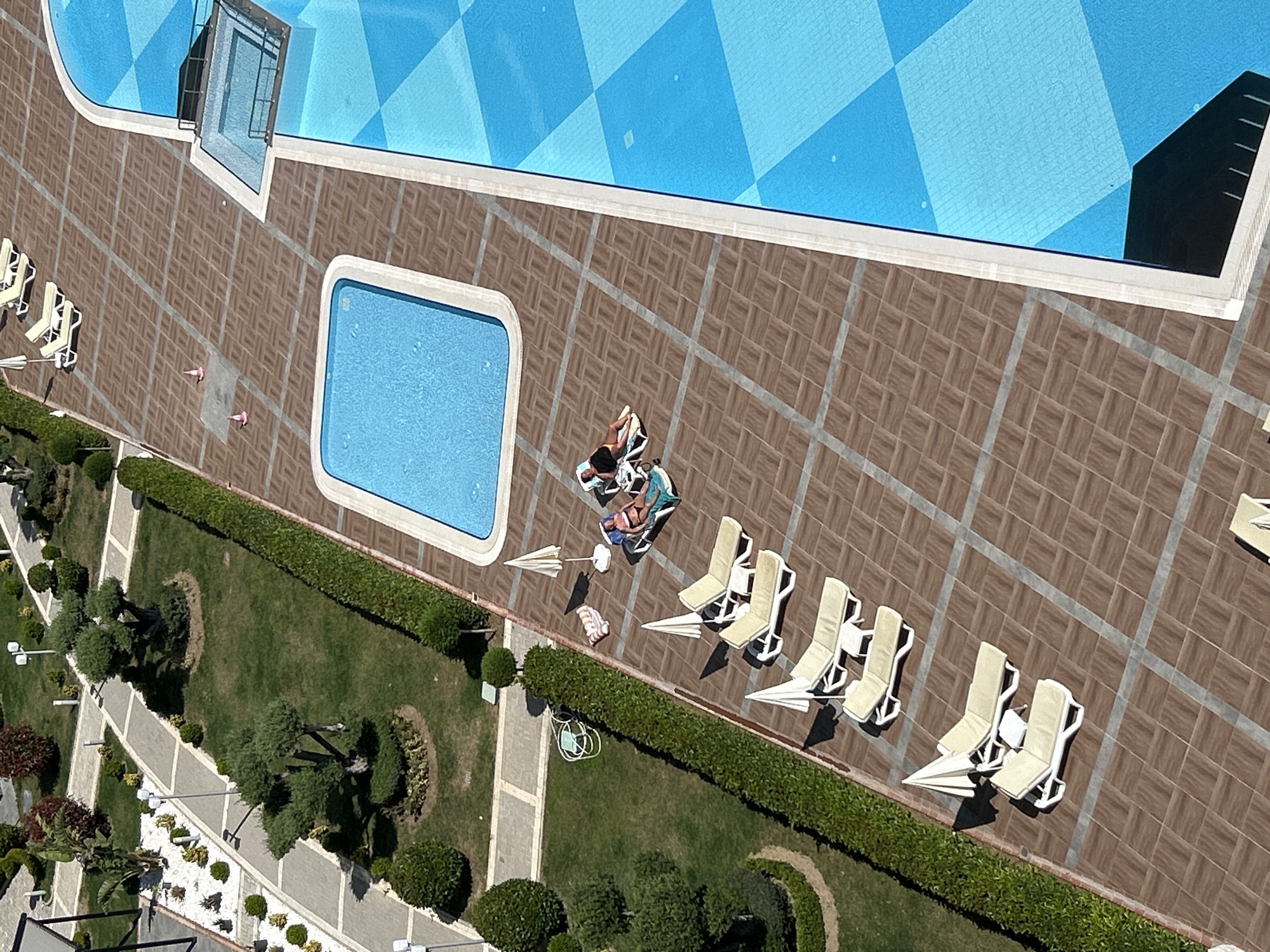 An aerial, high-angle shot captures a vibrant outdoor leisure area, likely a resort in Alanya, Turkey, on a clear, sunny day. The scene features two swimming pools: a large, irregularly shaped main pool with a distinctive blue and white checkered pattern on its bottom, and a smaller, rectangular wading pool with uniform blue water. Both pools are surrounded by a spacious sun deck paved with brown and grey geometric tiles.
Numerous light-colored lounge chairs with white frames are arranged on the tiled area. Several are accompanied by small, folded white sun umbrellas or larger, open white parasols, casting distinct shadows that suggest a mid-day sun.
At least three individuals are visible. Two people are relaxing on lounge chairs beside the smaller pool; one is sunbathing in dark swimwear, while the other is partially covered by a towel. Another person, wearing light pink attire, is seen standing or walking near the edge of the large pool. The overall activity appears to be casual relaxation and sunbathing.
Adjacent to the paved pool area is a meticulously maintained green garden, traversed by light-colored paved pathways. The garden is landscaped with various shrubs, small trees (some appearing to be olive trees with white gravel surrounds), and subtle outdoor lighting fixtures. A coiled garden hose is also visible in this area. A glass structure, possibly an access point or feature, extends into the main pool, and the shadow of a building is cast onto the water in the upper right, hinting at nearby architecture. No visible text is present in the image.