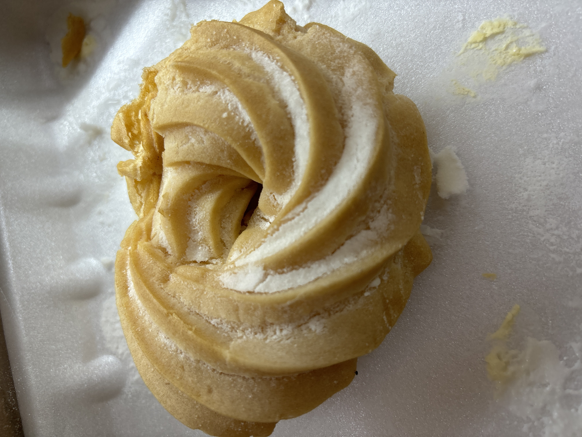 A single, golden-brown choux pastry, likely a cream puff shell, is centrally featured, resting on a white, textured surface, which appears to be a disposable foam container. The pastry exhibits a distinctive spiral or ruffled design, forming a ring with a central opening, and is lightly dusted with white powdered sugar, which accentuates its intricate ridges.
The white surface around the pastry shows scattered light yellow crumbs or residue, possibly from a cream filling, along with smaller white specks, likely additional powdered sugar or pastry fragments. The background is out of focus but consistent with the white container material. The lighting is even, suggesting an indoor setting.
No people are visible, and no specific activity or event is depicted, beyond the presentation of the pastry. There is no visible text. The image provides a close-up view of the dessert, highlighting its texture and details. While the context of Malki Iskar, Bulgaria, is provided, there are no visual cues within the image itself to indicate this location.